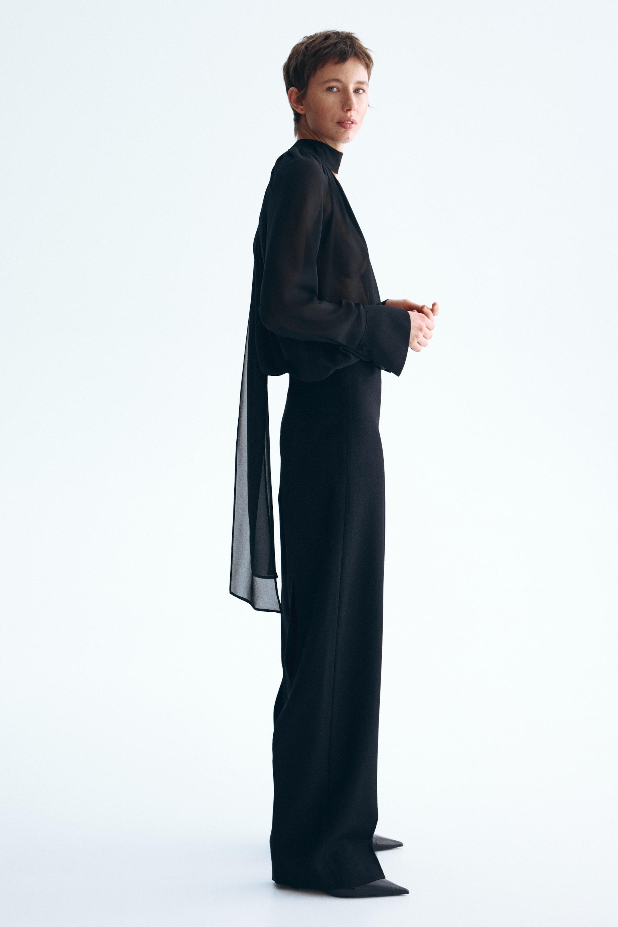 View larger image: The model is donning an oversized black blouse crafted from a sheer weave. It showcases a scarf collar tied at the neck, with elongated ends cascading down the back, and long sleeves finished with buttoned cuffs.