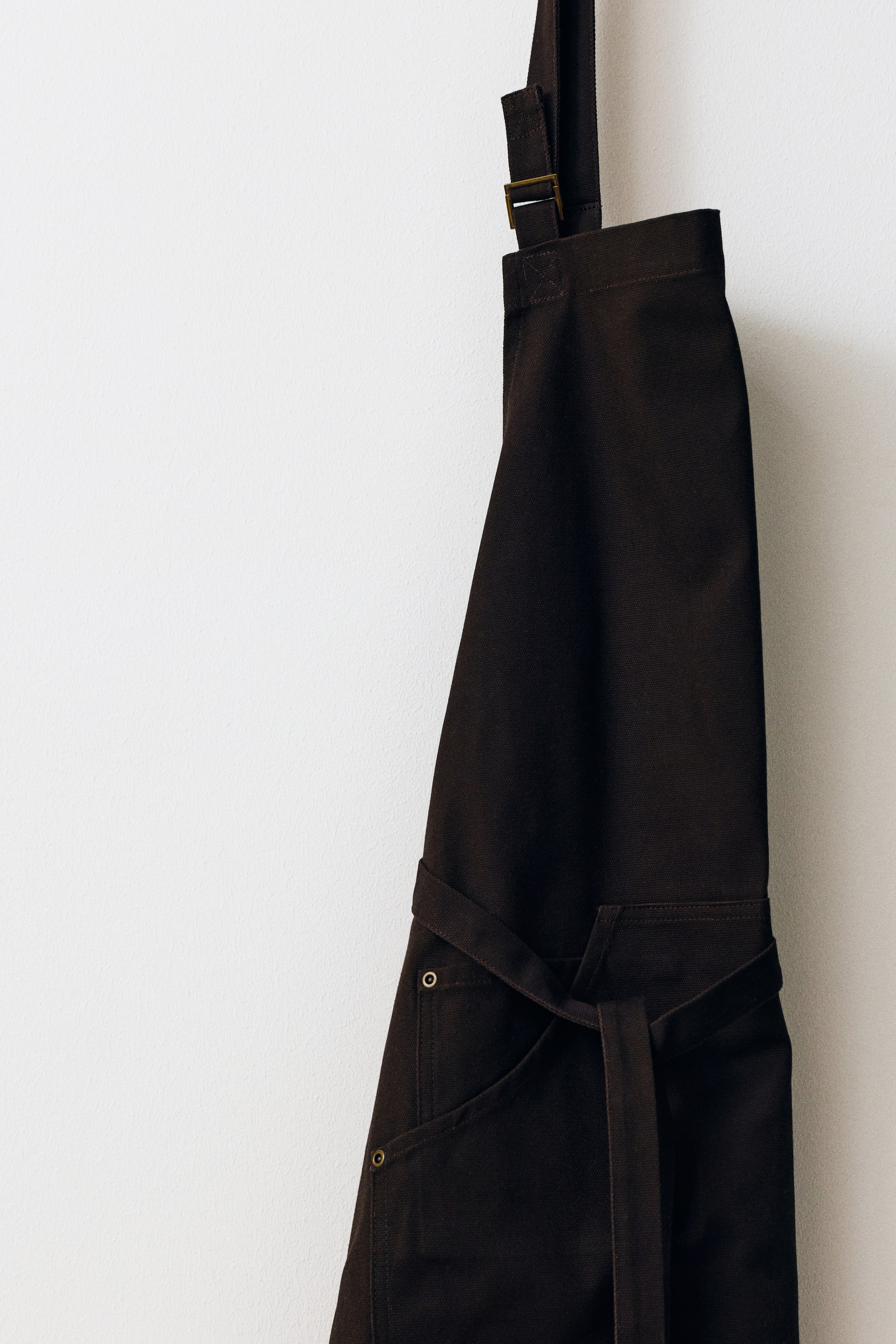 Voir une image plus grande: Un tablier en toile brun foncé est suspendu en vue de profil, doté d’une sangle de cou réglable avec une boucle métallique et d’une ceinture à nouer enroulée à l’avant. Le tablier présente une grande poche à l’avant, renforcée par de petits rivets métalliques aux coins.