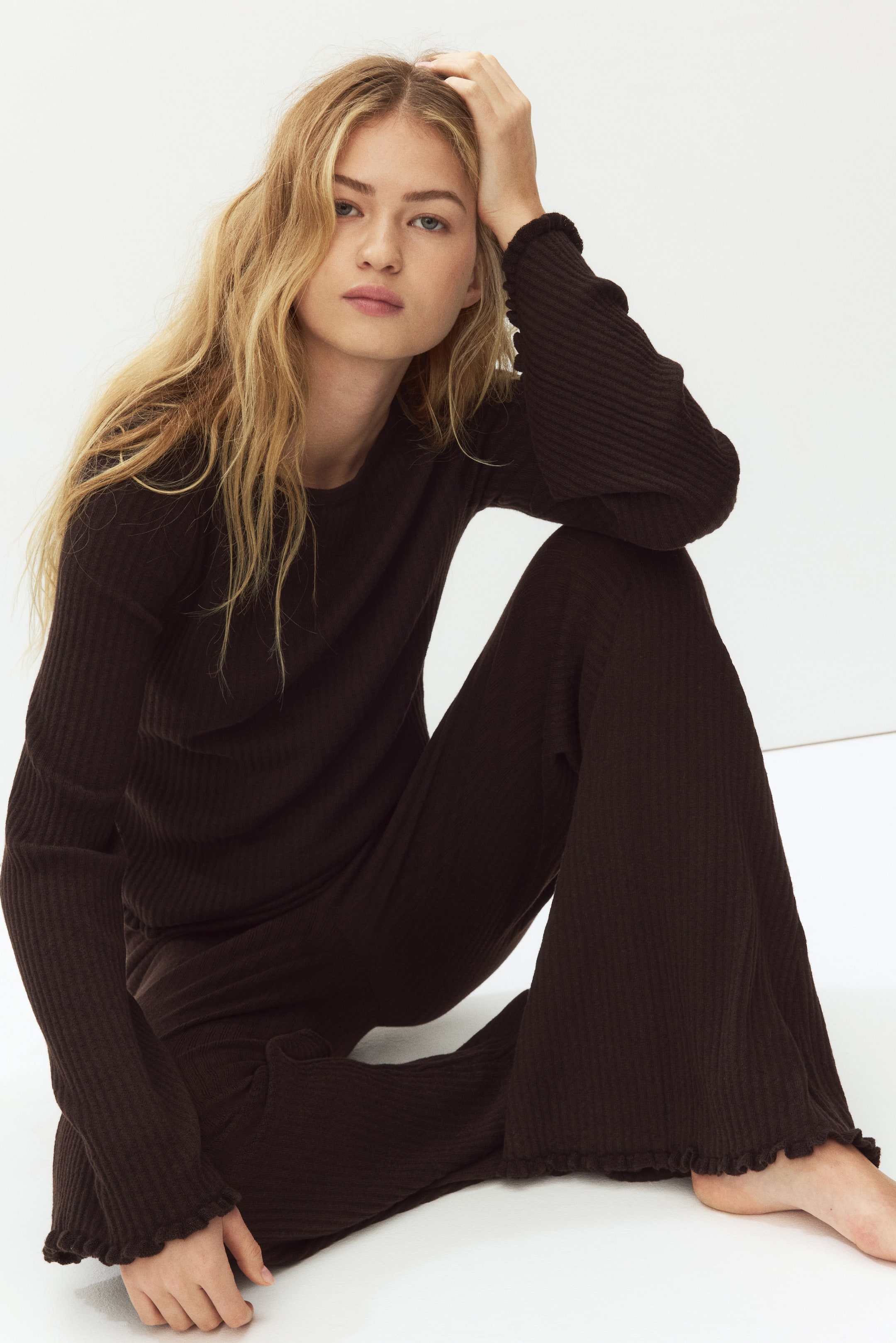 View larger image: Wearing a relaxed fit dark brown ribbed loungewear set, featuring a long-sleeved crew neck top and wide-leg pants, with lettuce edges finishing the top's cuffs and pant hems.