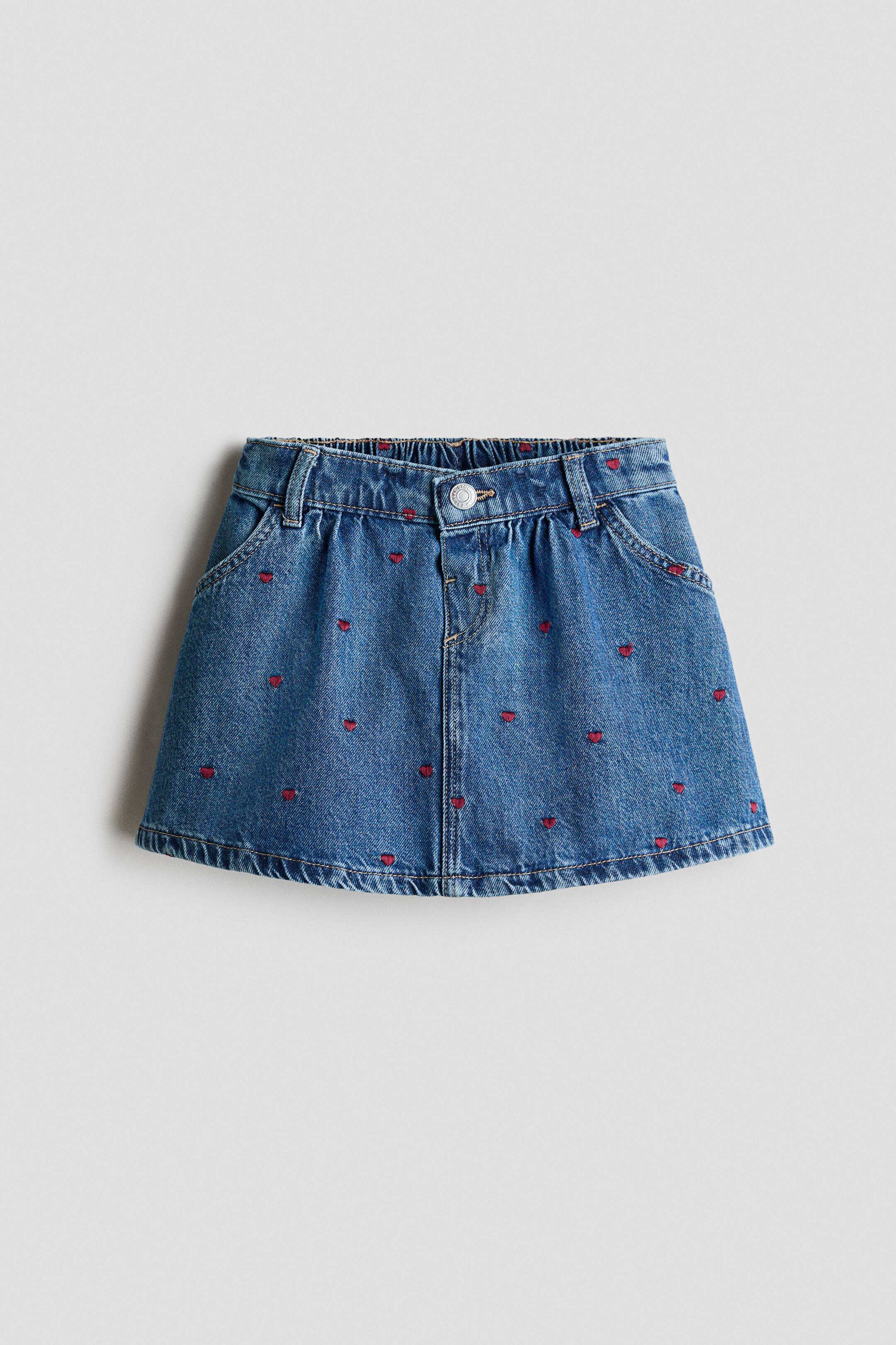 View larger image: A blue denim skirt, front-facing, featuring an A-line silhouette with an all-over pattern of small, embroidered red hearts. The skirt features gathered detailing along the waistband, a center button closure, and curved side pockets.