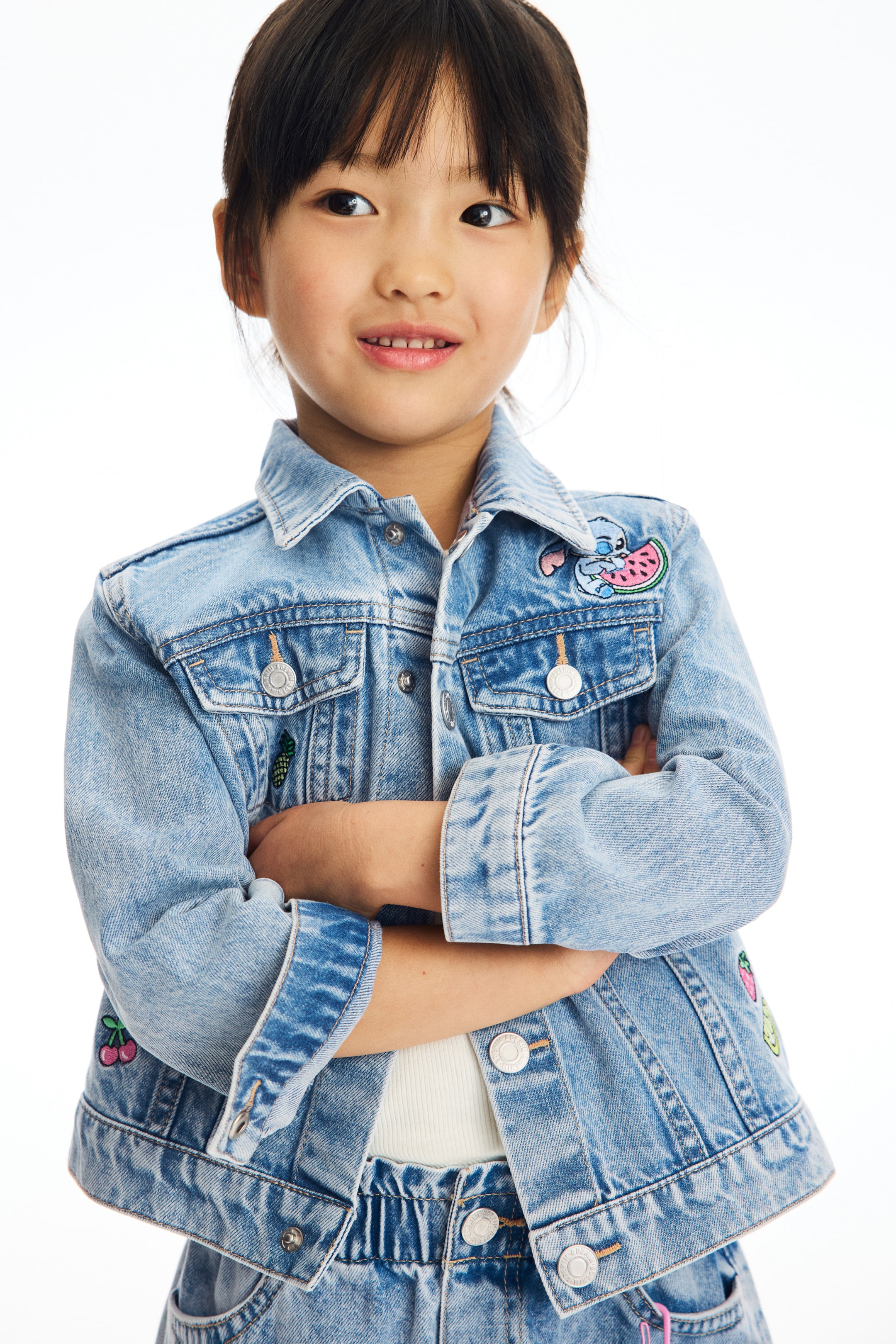 View larger image: A light blue denim jacket is showcased from a front view, featuring a classic pointed collar, silver-toned button front closure, and two buttoned chest flap pockets. The jacket is embellished with several colourful patches, including a blue character holding a watermelon, a pineapple, and cherries.