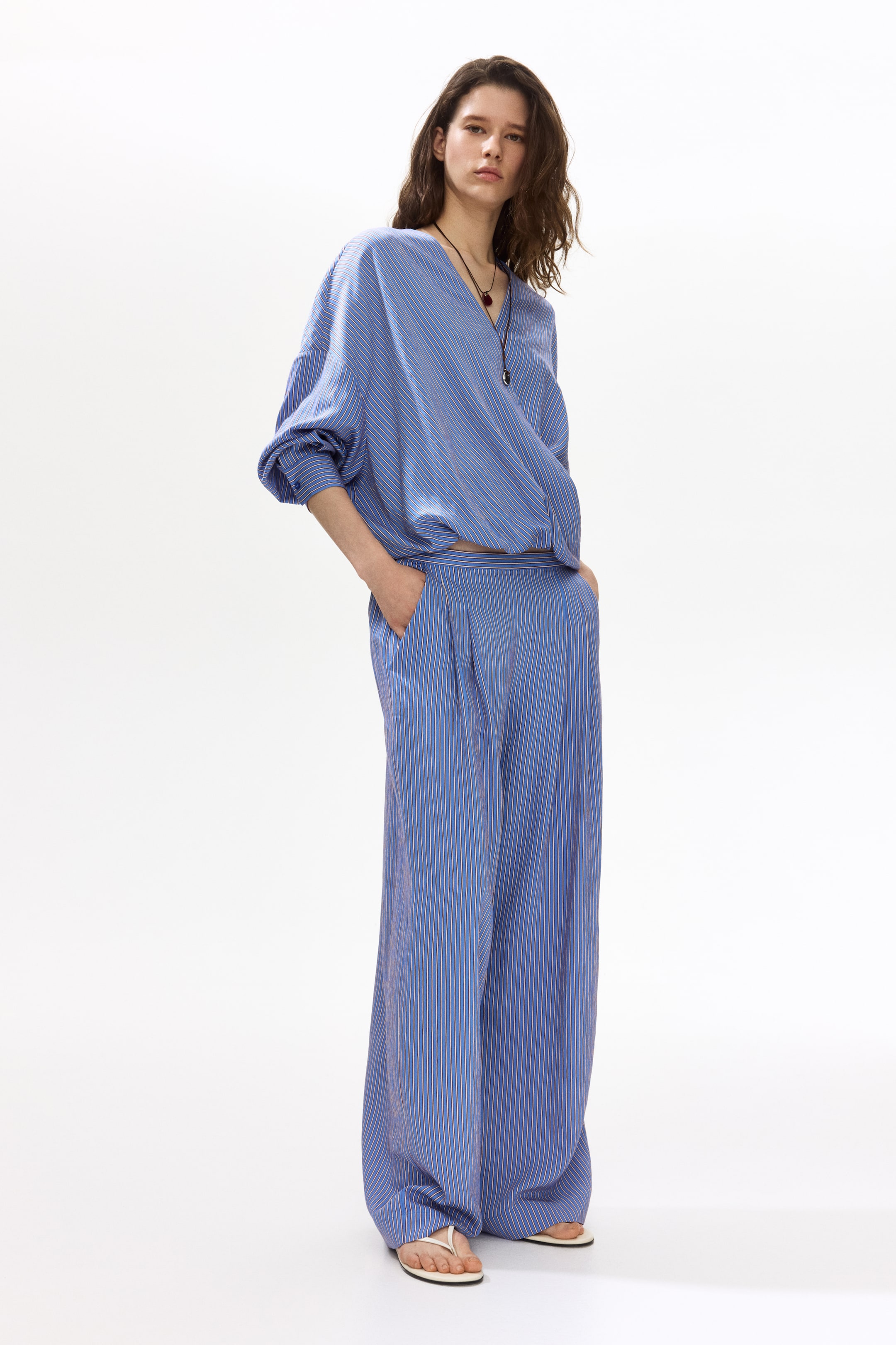 View larger image: The blue and white pinstriped two-piece set features a front-facing long-sleeved wrap top with a V-neck, voluminous sleeves, and a gathered hem. It pairs with pleated wide-leg trousers that include side pockets.