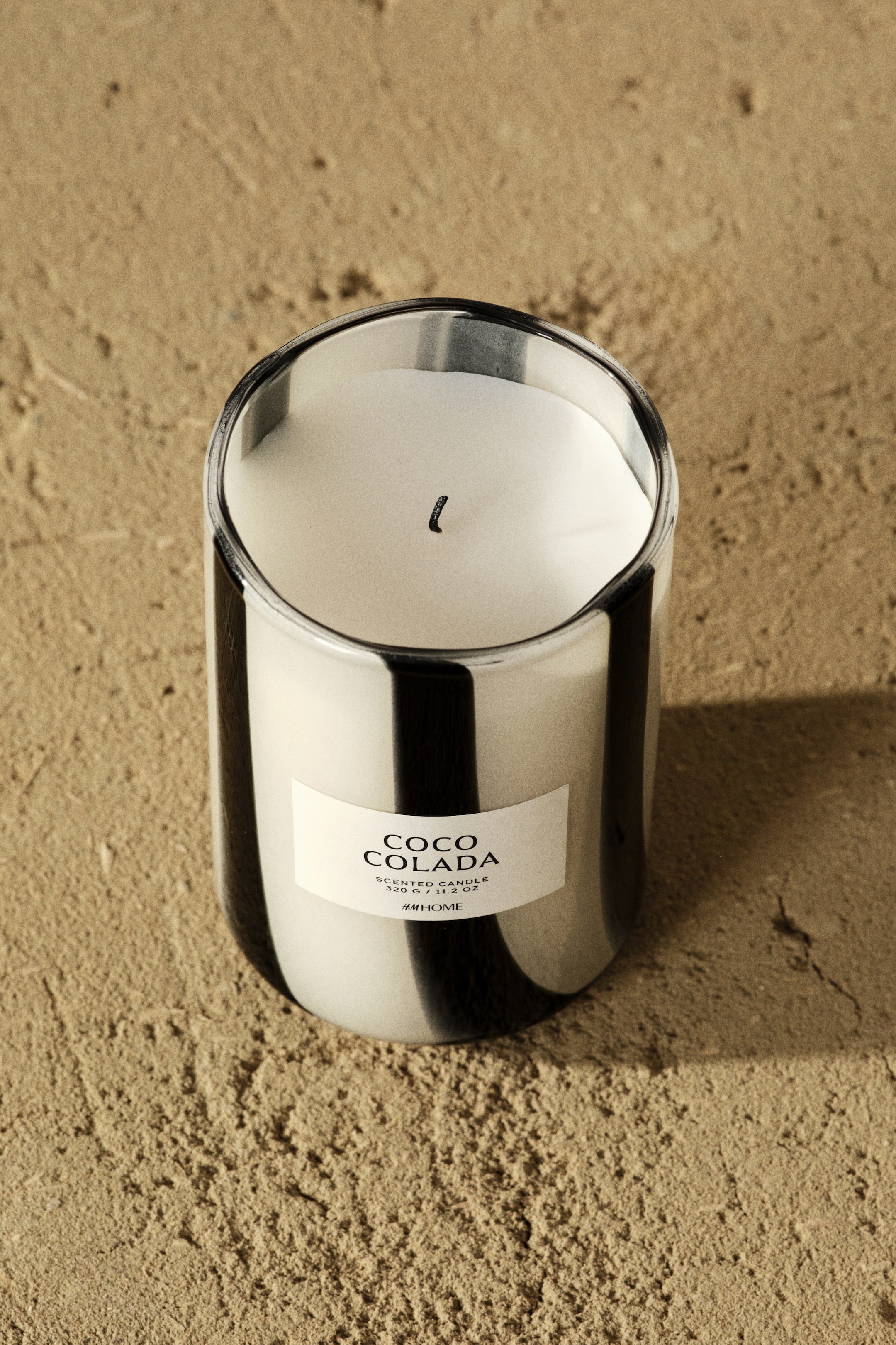 Vis større bilde: En sylindrisk duftlys står oppreist, sett fra en høy vinkel, med sitt glasskrukke som har svarte vertikale striper på en metallisk overflate. Krukken er fylt med hvit voks, med en svart veke som stikker ut fra midten, og en rektangulær etikett med "COCO COLADA" er festet på forsiden.