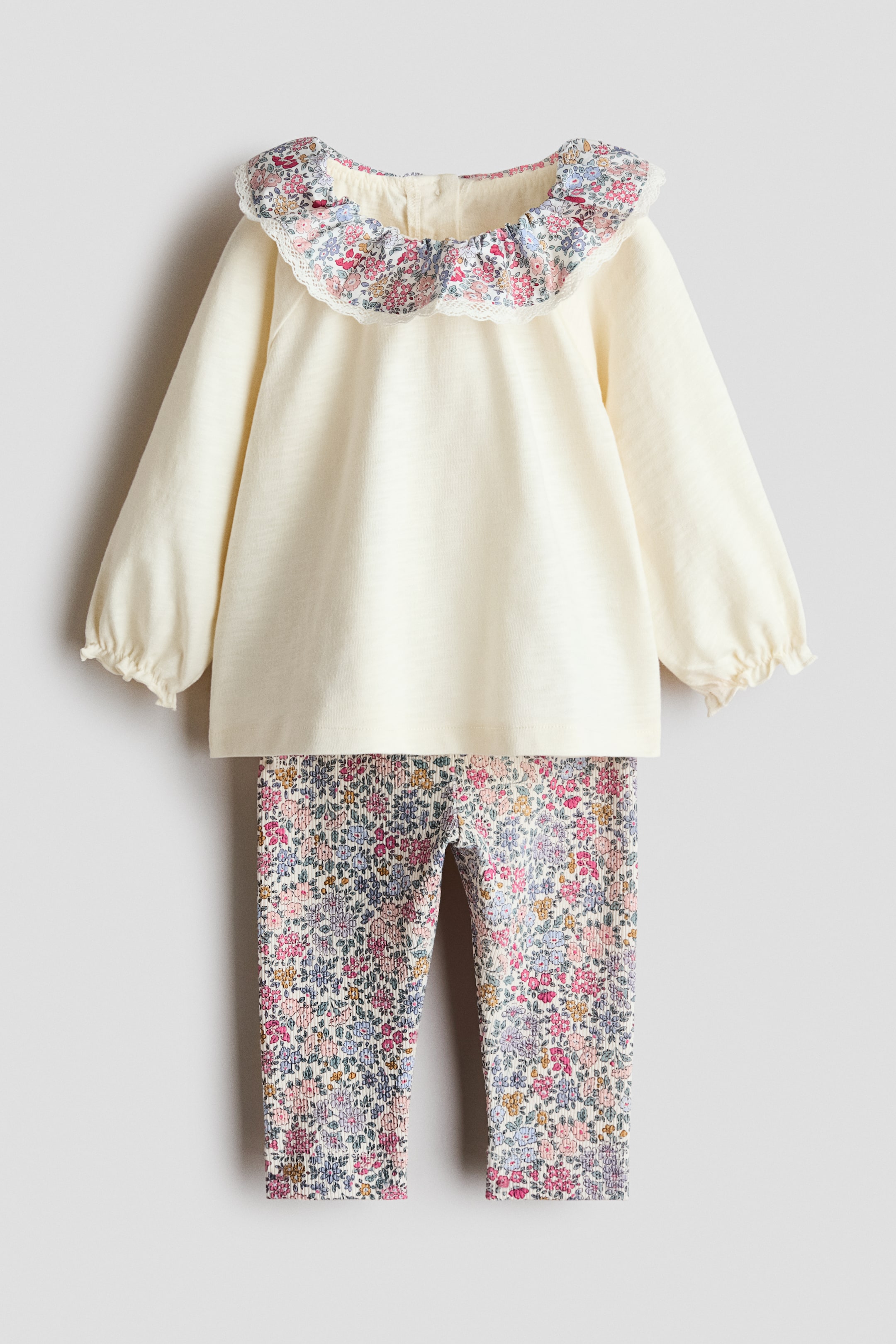 Voir une image plus grande: Un ensemble pour bébé en deux pièces vu de face présente un haut crème à manches longues avec un col à volants floral, bordé de dentelle blanche. Les poignets du haut sont froncés avec un élastique, et un legging en maille côtelée à imprimé floral assorti apparaît en dessous.