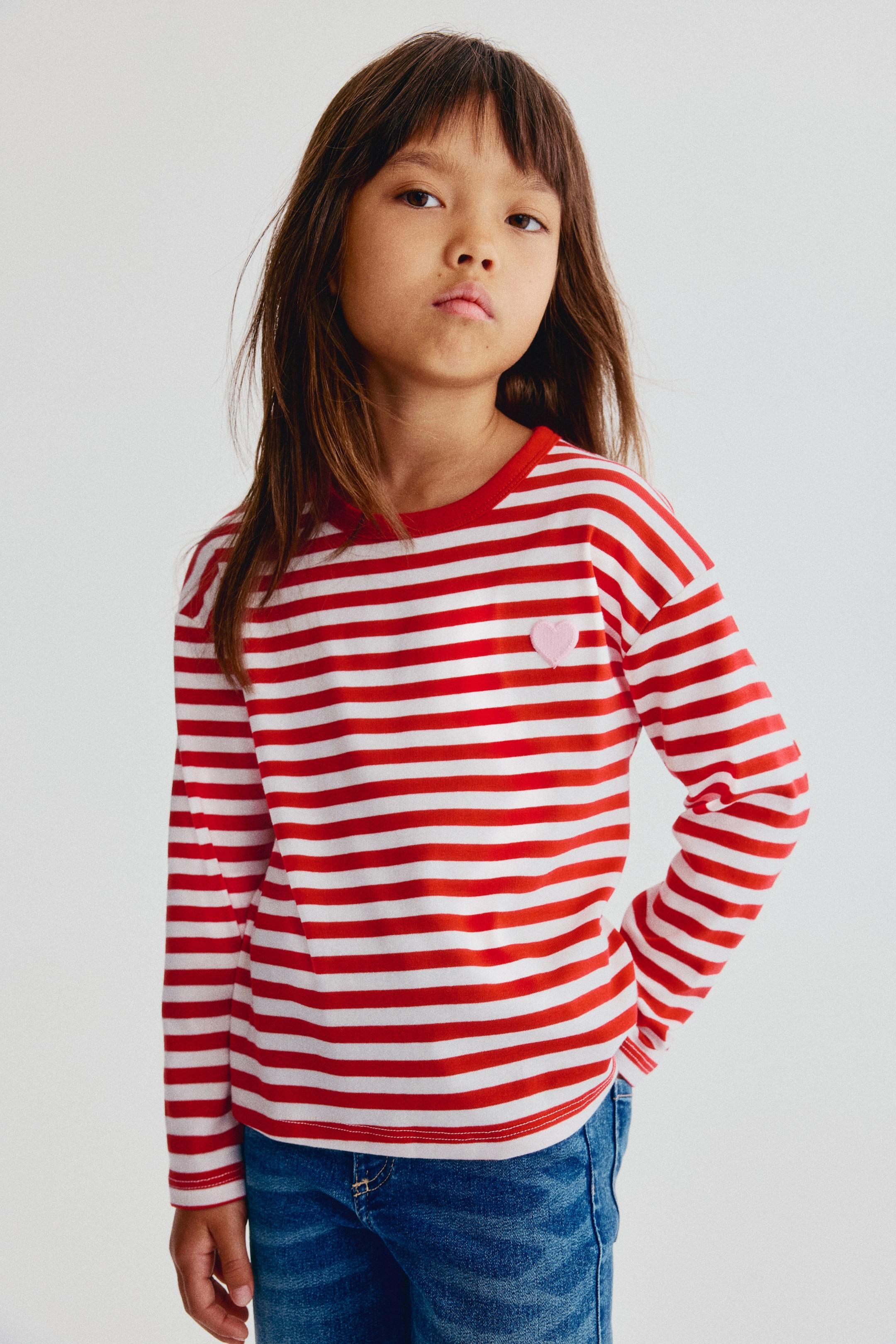 View larger image: A long-sleeve T-shirt featuring red and white horizontal stripes is shown from the front, showcasing a round, red ribbed neckline, dropped shoulders, and a small pink embroidered heart on the left chest. The T-shirt is styled with blue denim jeans.