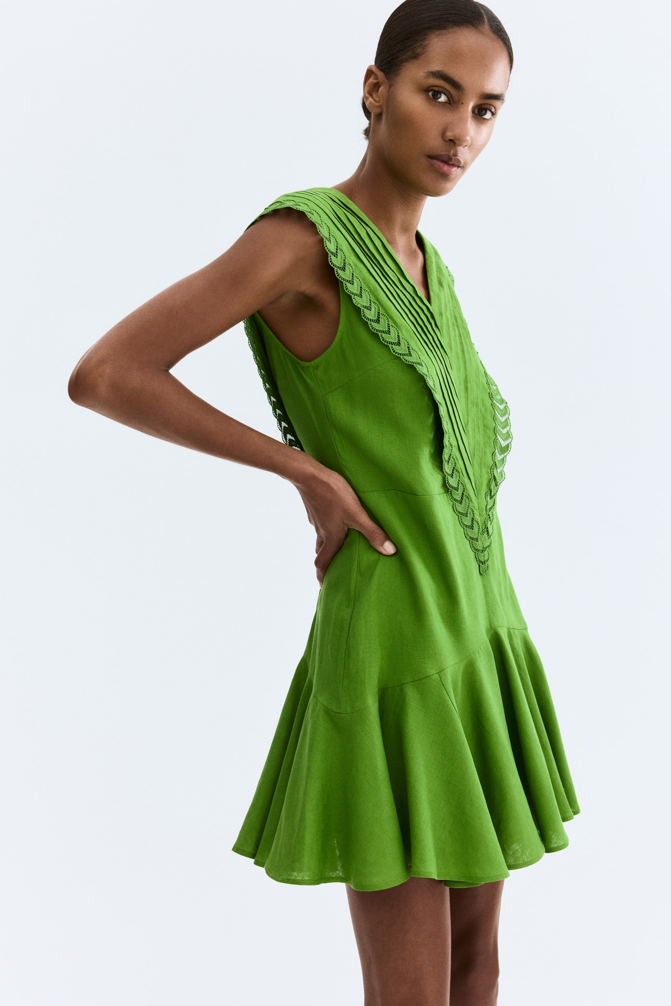 Ampliar la imagen: Un vestido verde sin mangas de corte mini presenta un escote en V con pliegues verticales en el frente del corpiño. Un ribete trenzado con bordes festoneados adorna los bordes del escote en V y las sisas, y la falda se ensancha en el dobladillo.