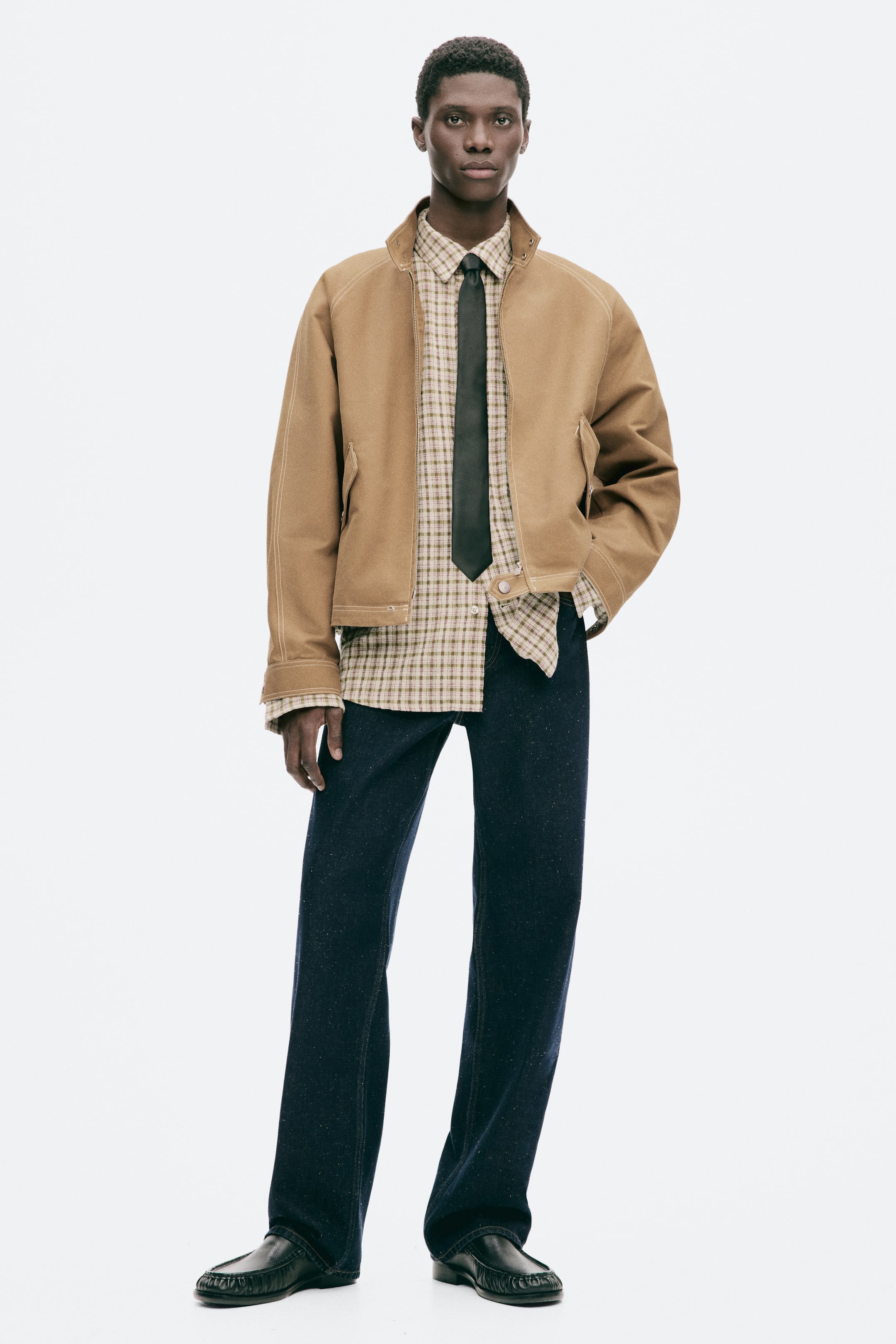 View larger image: A tan workwear jacket with visible contrast stitching is worn by the model, featuring a stand collar with snap buttons, raglan sleeves with buttoned cuffs, diagonal flap pockets with snap buttons, and a straight hem with a button tab at the front. The jacket is open, revealing a plaid shirt and tie, and the model is placing a hand in a pocket.