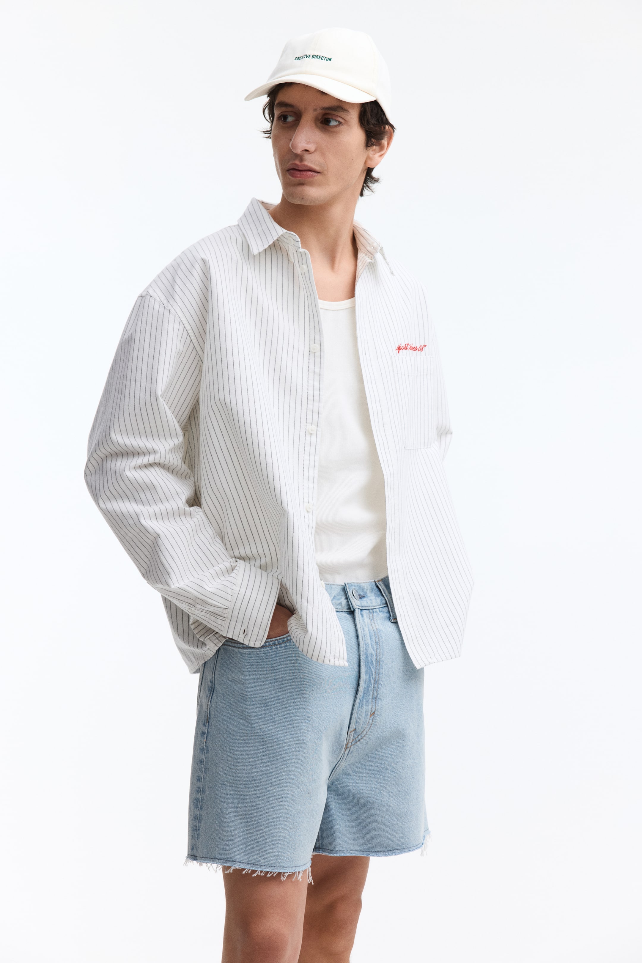 View larger image: The model is wearing light blue rigid cotton denim shorts with five pockets. These shorts have a baggy fit, a regular waist with a visible zip fly, and raw-edge hems. The model has one hand casually placed in a front pocket of the shorts.