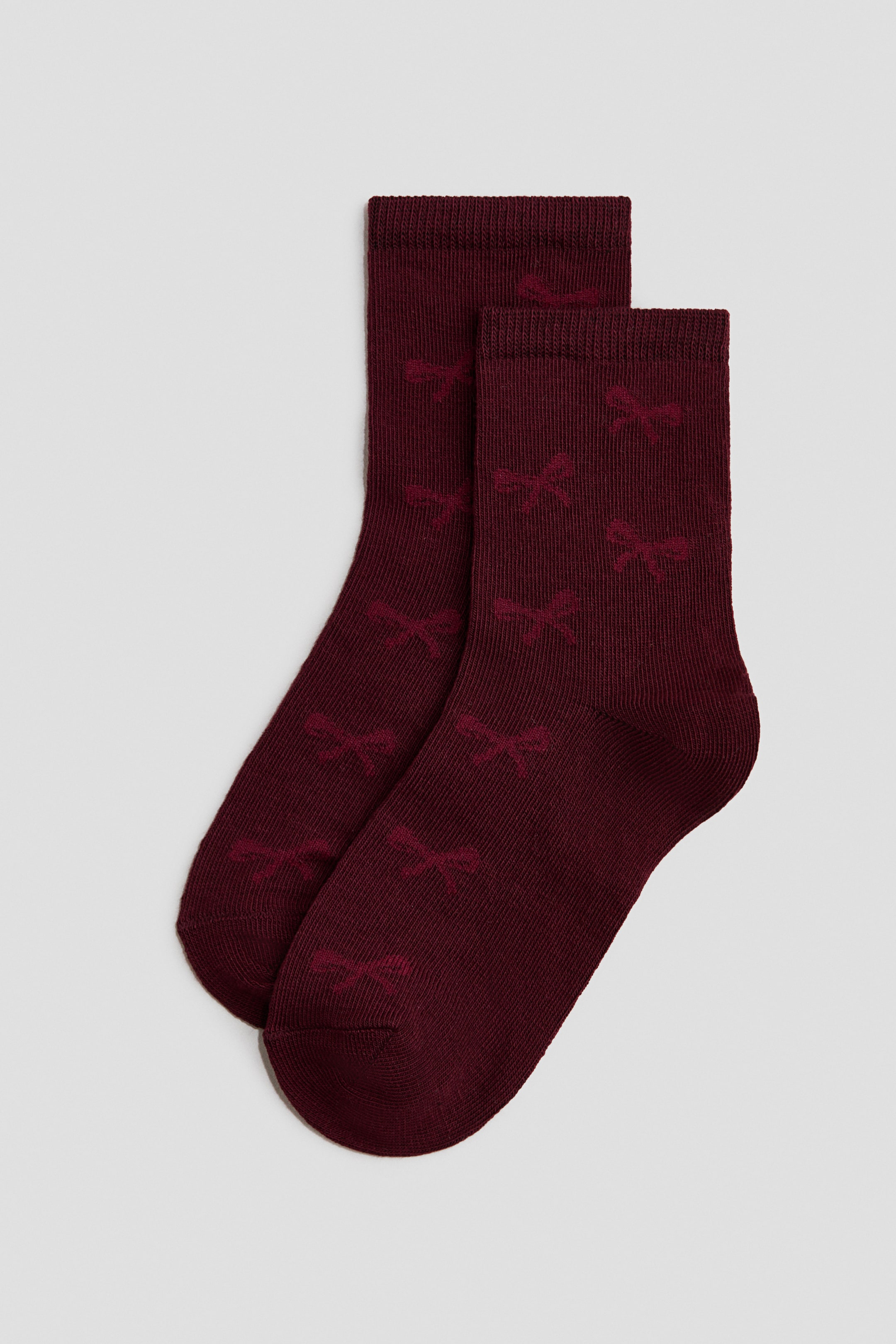 Agrandir l'image: Une paire de chaussettes arbore une couleur bordeaux profond avec un motif répétitif de nœuds rouges plus foncés et subtils. Elles présentent des poignets côtelés en haut et sont exposées de FACE, à plat, l'une chevauchant légèrement l'autre.