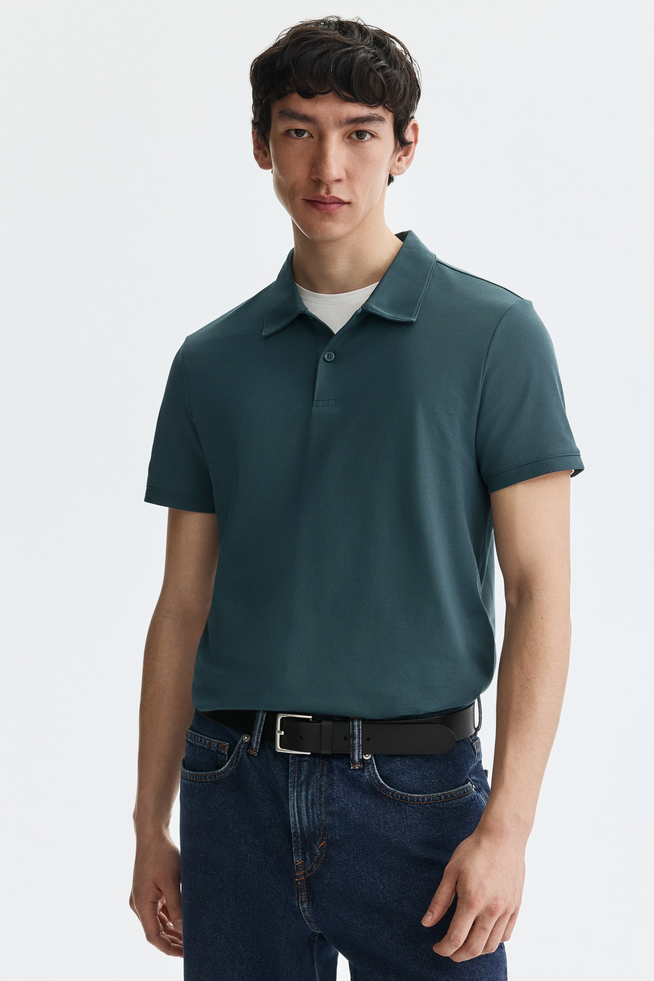 Voir une image plus grande: Un polo bleu sarcelle foncé est présenté de face, confectionné dans un tissu tricoté lisse, avec des manches courtes, un col classique et une patte de boutonnage à deux boutons, laissant entrevoir un t-shirt à col rond blanc superposé en dessous.
