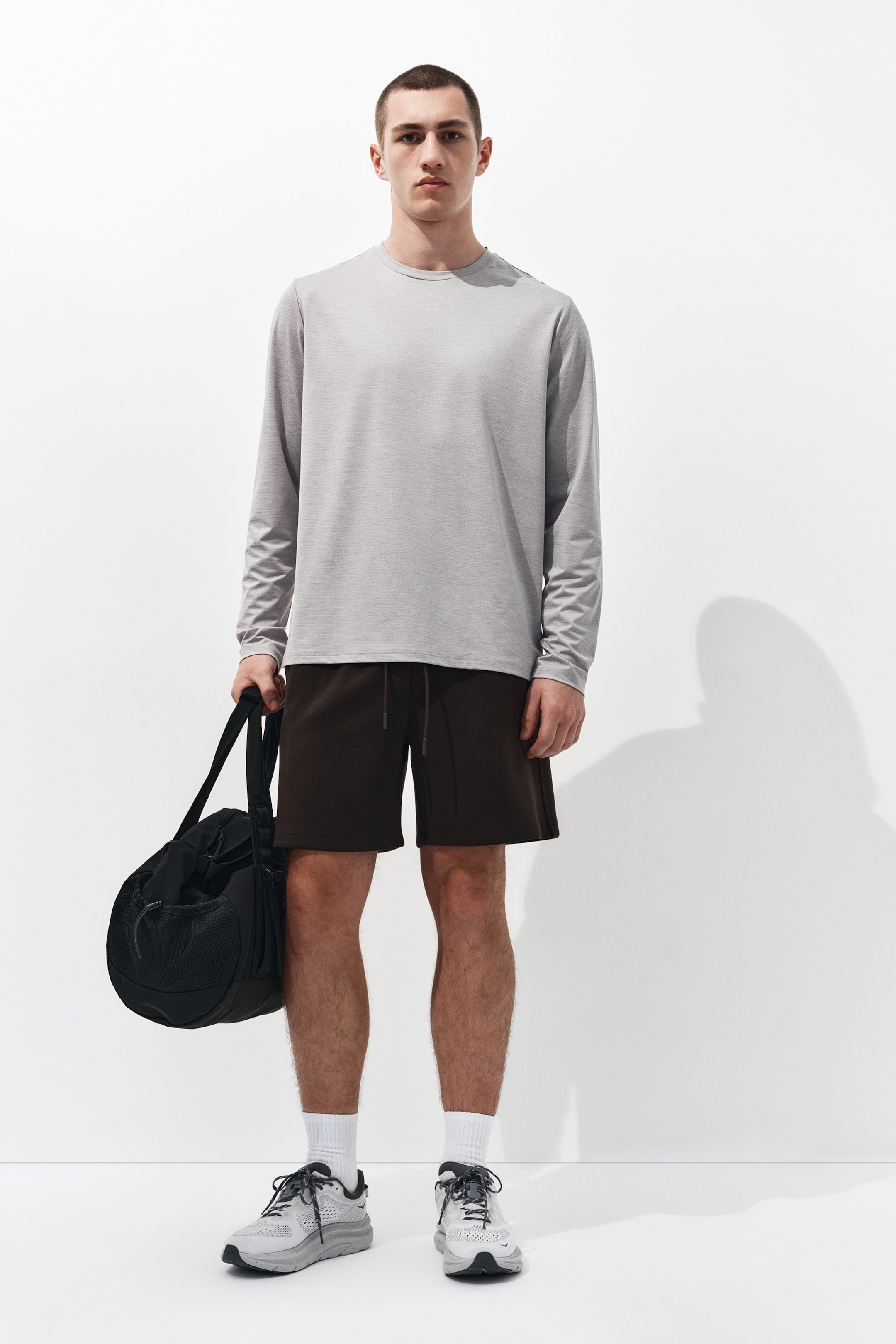 Ampliar la imagen: Se está usando una camiseta de manga larga con cuello redondo de color gris jaspeado claro, que presenta un ajuste relajado. Unos shorts deportivos marrón oscuro con una cintura de cordón visible complementan la camiseta, cayendo justo por encima de la rodilla.