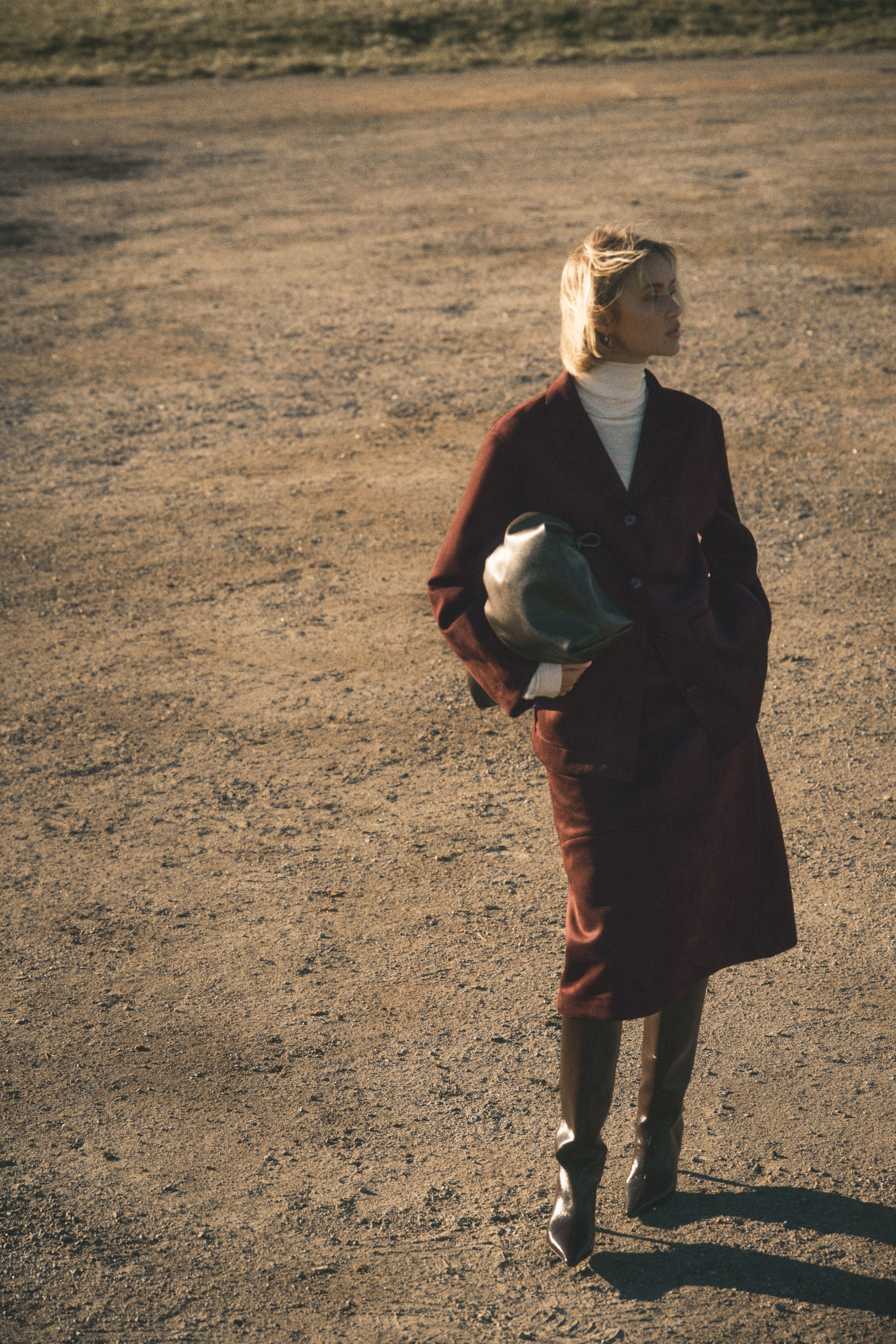 查看大圖: 酒紅色單排扣西裝外套，帶有平駁領，搭配一條配套的及膝裙，展示在一個稍微向右側傾斜的人偶上。這件外套下面穿著一件奶油色的高領毛衣，而高且暗色的尖頭靴子完成了整體搭配，同時還拿著一個深色的柔軟結構的包包。