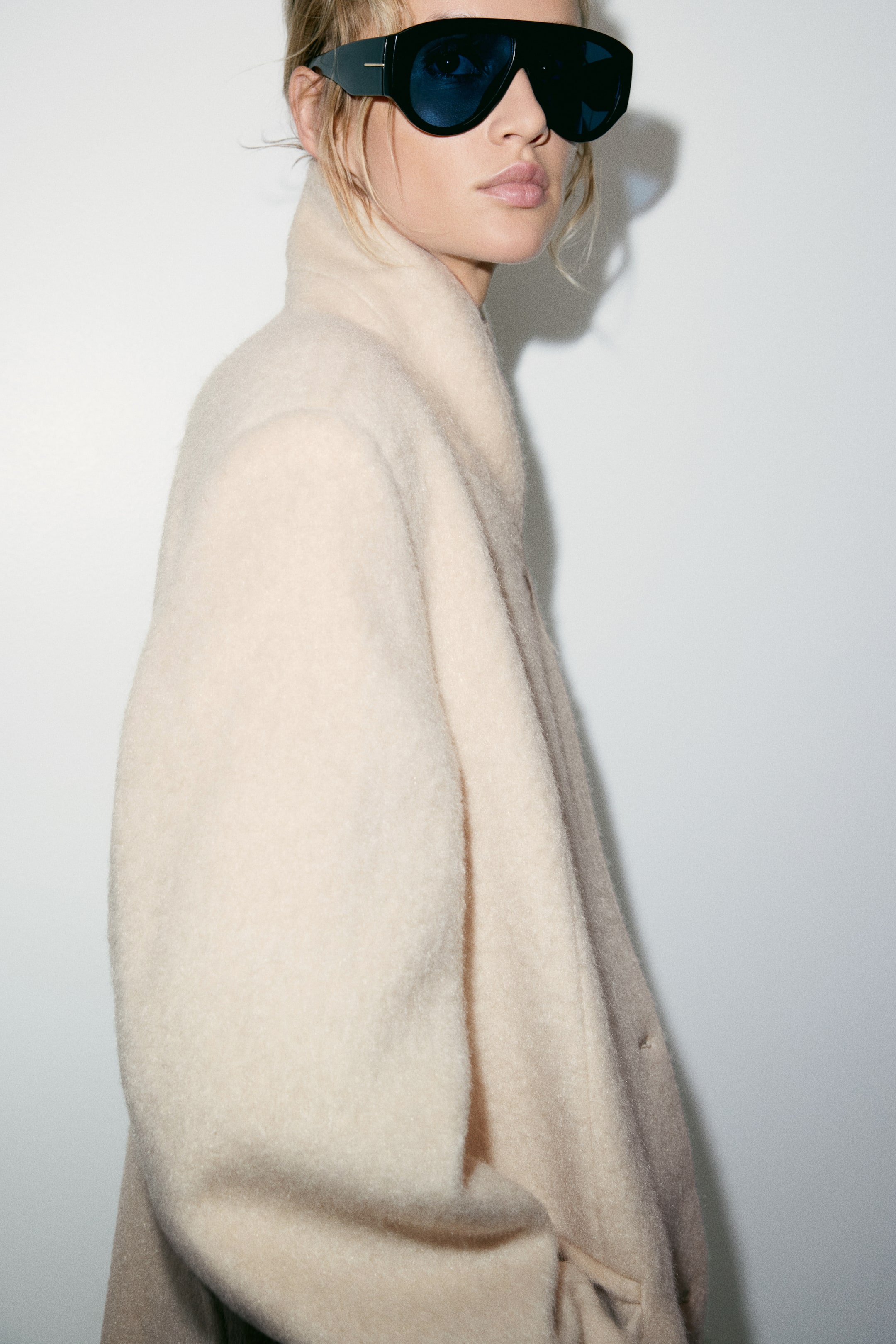 Zobraziť väčší obrázok: Model má na sebe oversized kabát v krémovej farbe z vlnenej zmesi, ktorý zvýrazňuje jeho mäkkú plstenú štruktúru a vysoký golier. Kabát je doplnený veľkými čiernymi štítovými slnečnými okuliarmi.