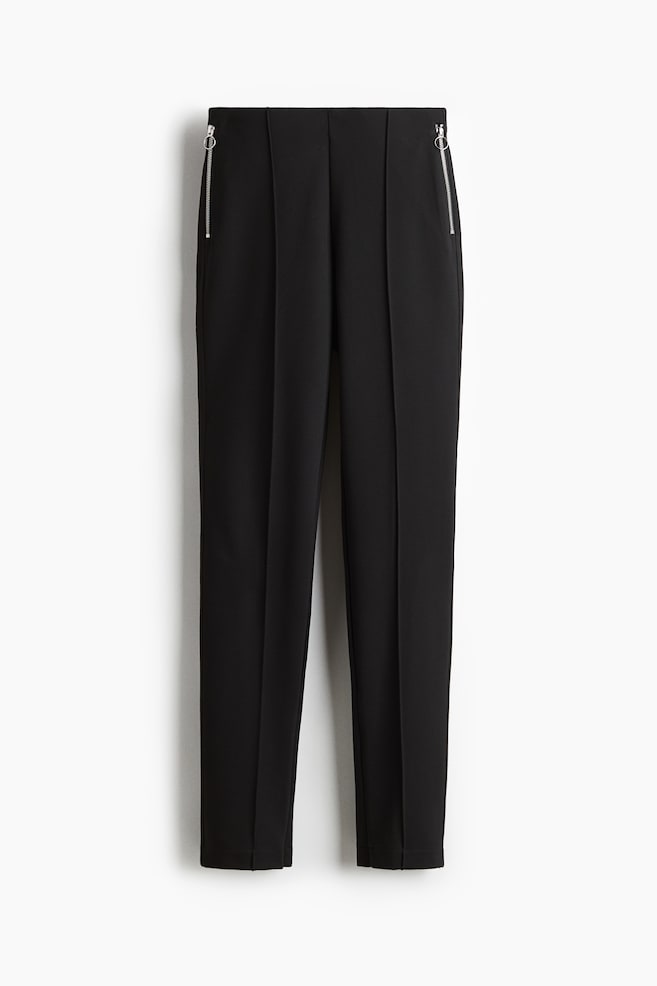 Pantalones negros de corte ajustado presentan su parte delantera, con una cintura alta y bolsillos laterales con cremallera en tono plateado, además de pliegues cosidos que recorren la parte delantera de cada pierna. - 1