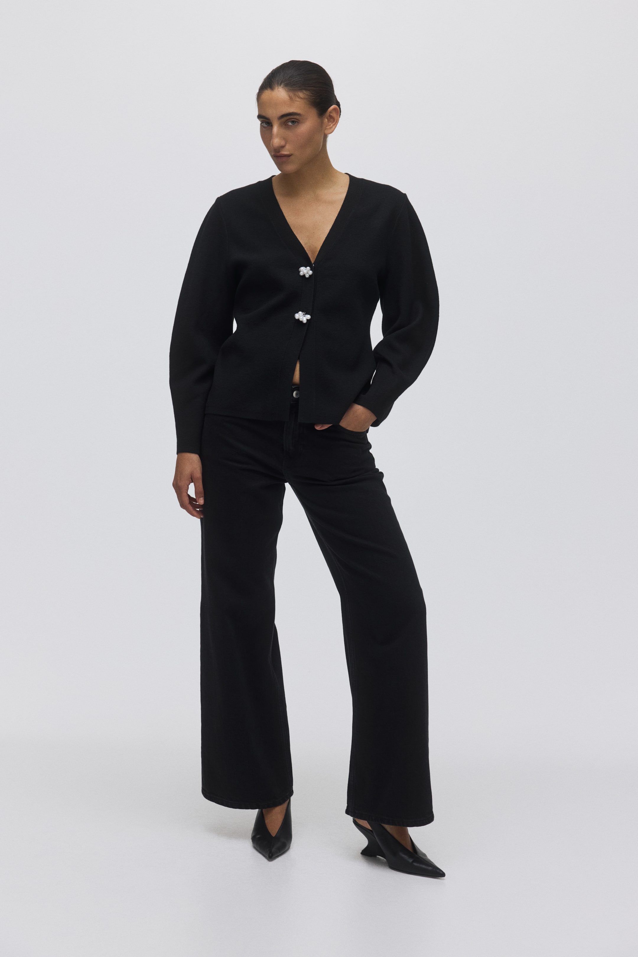 View larger image: The black V-neck cardigan is facing front, featuring long, subtly voluminous sleeves and a straight hem ending at the waist. It is fastening at the front with two light-coloured, clustered buttons.