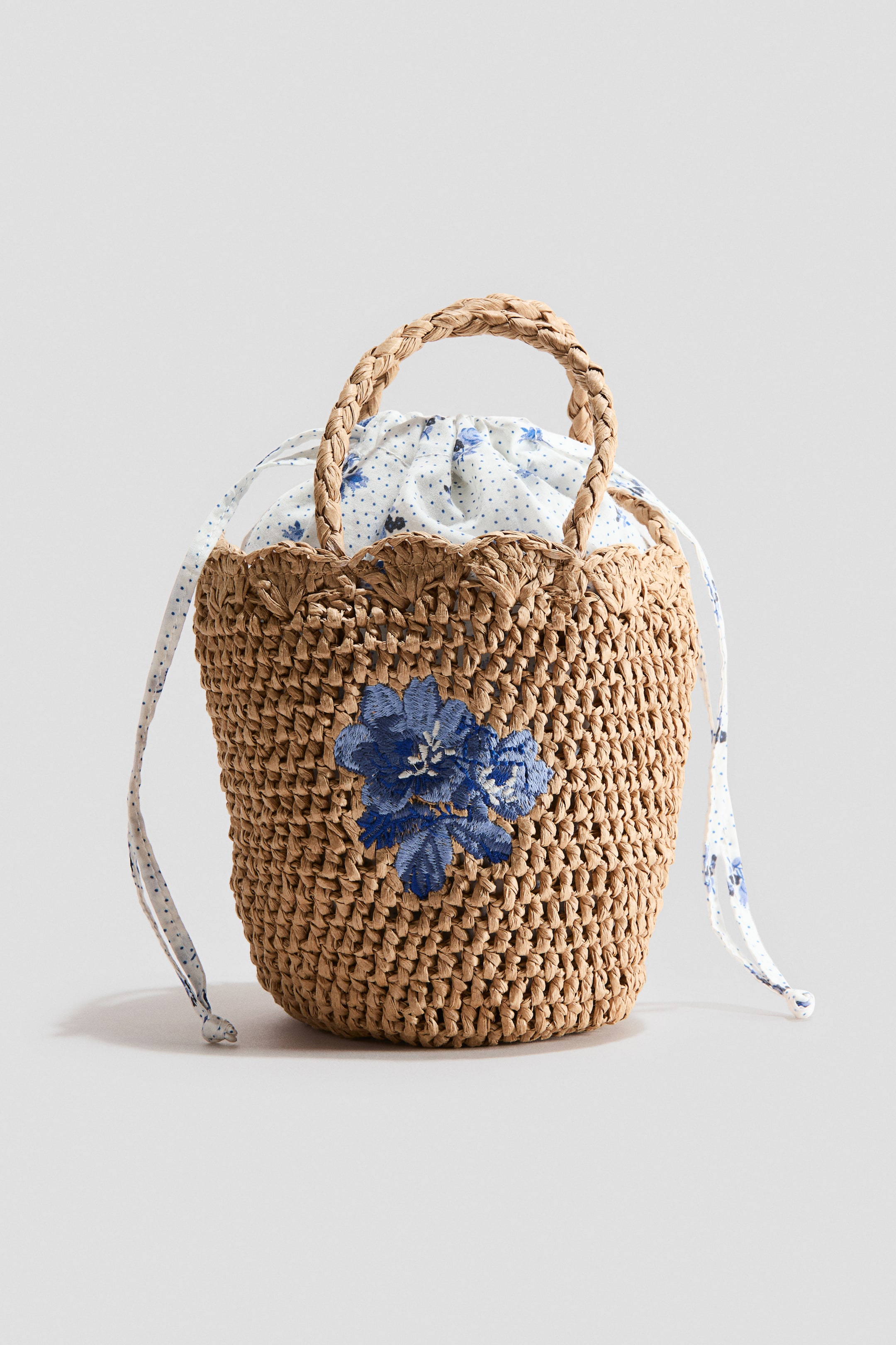 Grotere afbeelding bekijken: Een strooien emmer tas is geweven met een bruine strooien body en heeft blauwe bloemenborduursels aan de voorkant. Een voering van wit en blauw bloemenpatroon piept eruit, vormt een geschulpte rand en sluit met een bijpassend trekkoord, terwijl een gevlochten strooien handvat over de bovenkant buigt.