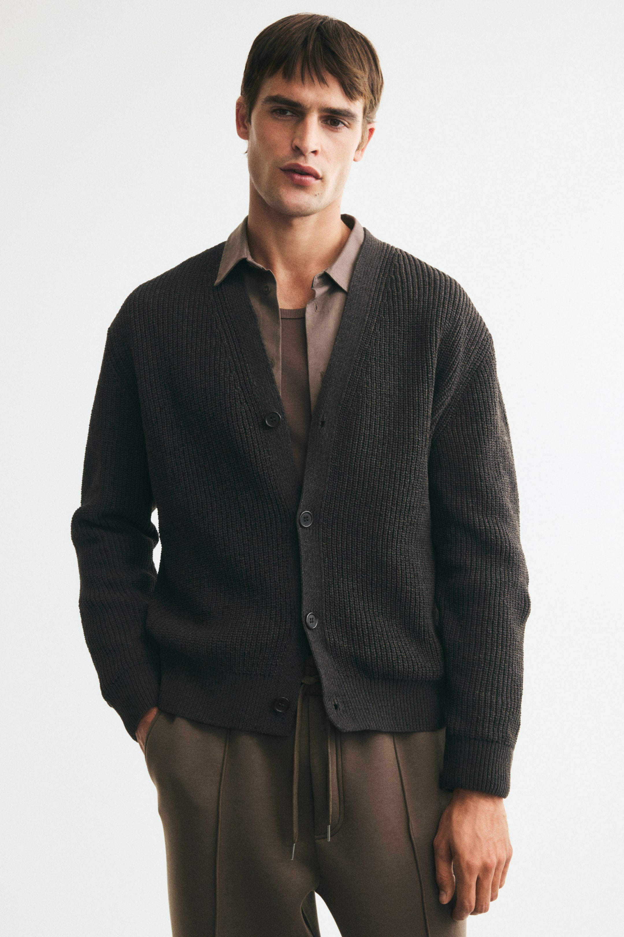 View larger image: A dark grey ribbed cardigan, featuring a V-neckline, dropped shoulders, and buttons down the front, is worn open by the model over a brown collared shirt and a darker brown T-shirt. The model is resting one hand in the pocket of brown trousers, which are paired with the cardigan.