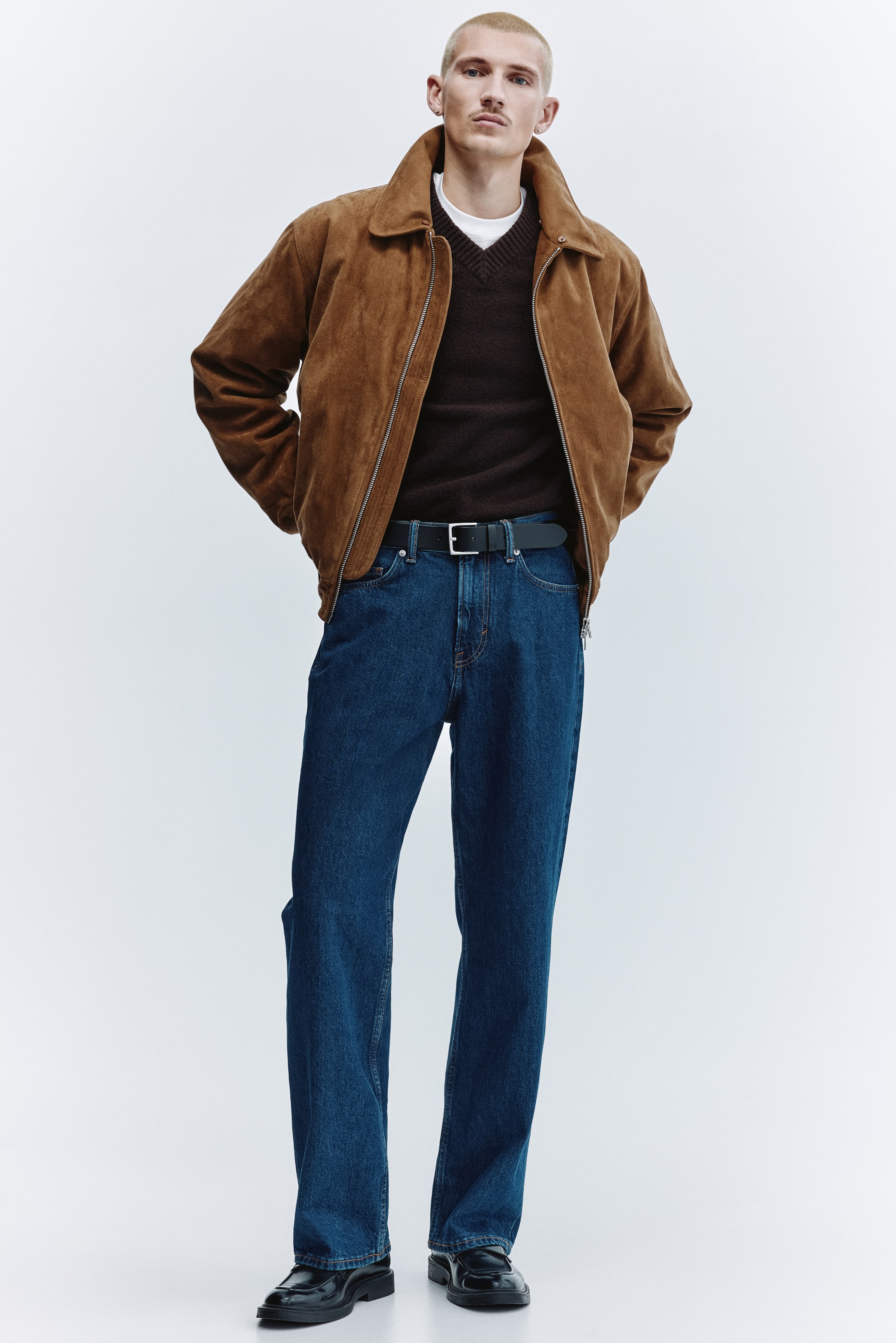 View larger image: The men's ensemble showcases a brown suede bomber jacket with a spread collar, layered over a dark brown V-neck sweater and a white T-shirt. Dark blue straight-leg jeans feature a black leather belt with a silver buckle and are styled with black chunky loafers.