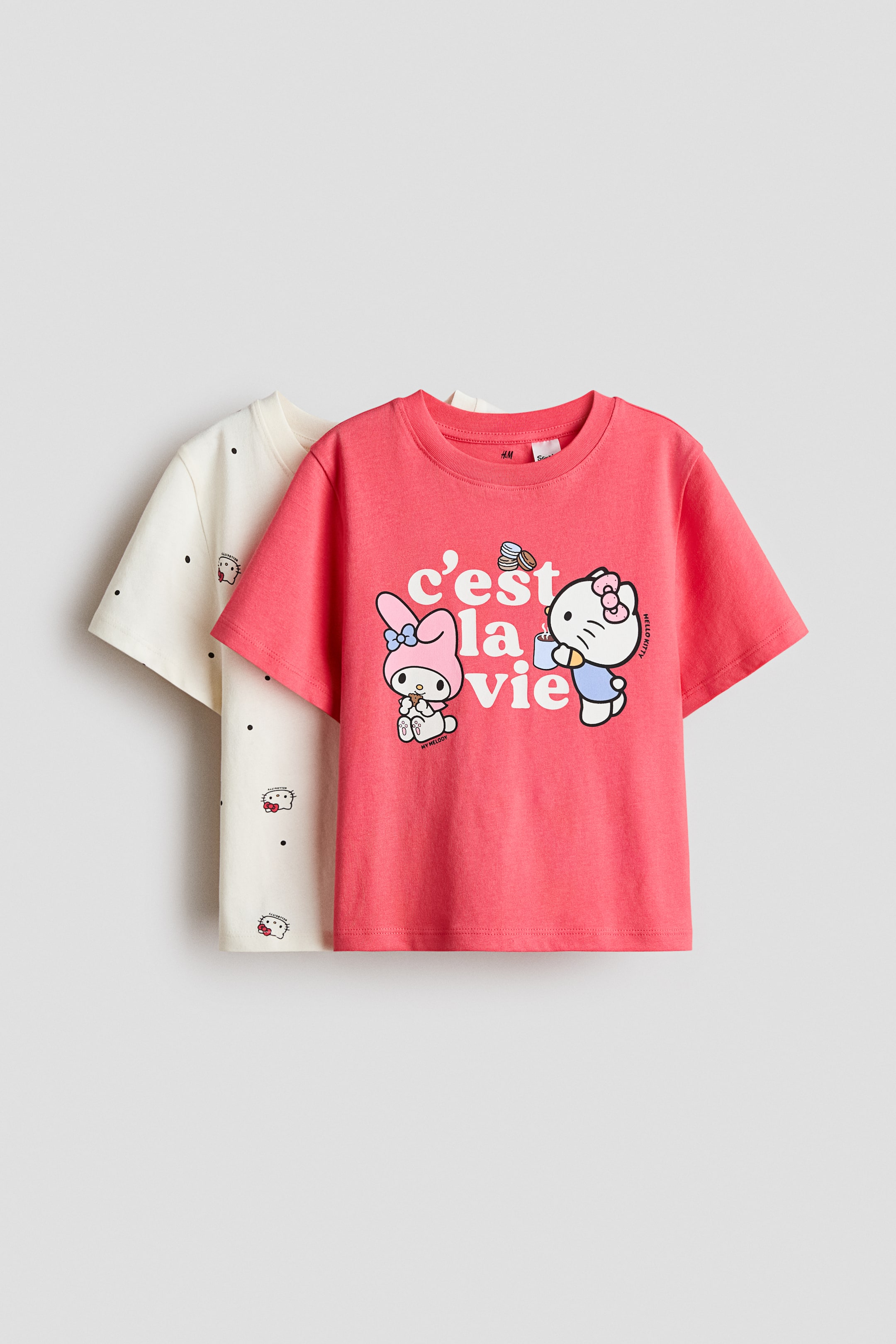 Größeres Bild ansehen: Zwei kurzärmelige T-Shirts mit Rundhalsausschnitt werden präsentiert. Das vordere T-Shirt in Korallenrosa zeigt einen Grafikdruck von My Melody und Hello Kitty mit dem Text "c'est la vie". Das zweite T-Shirt, das hinter dem ersten sichtbar ist, ist in Off-White gehalten und zeigt ein sich wiederholendes Muster von Hello Kitty-Gesichtern und kleinen schwarzen Punkten.