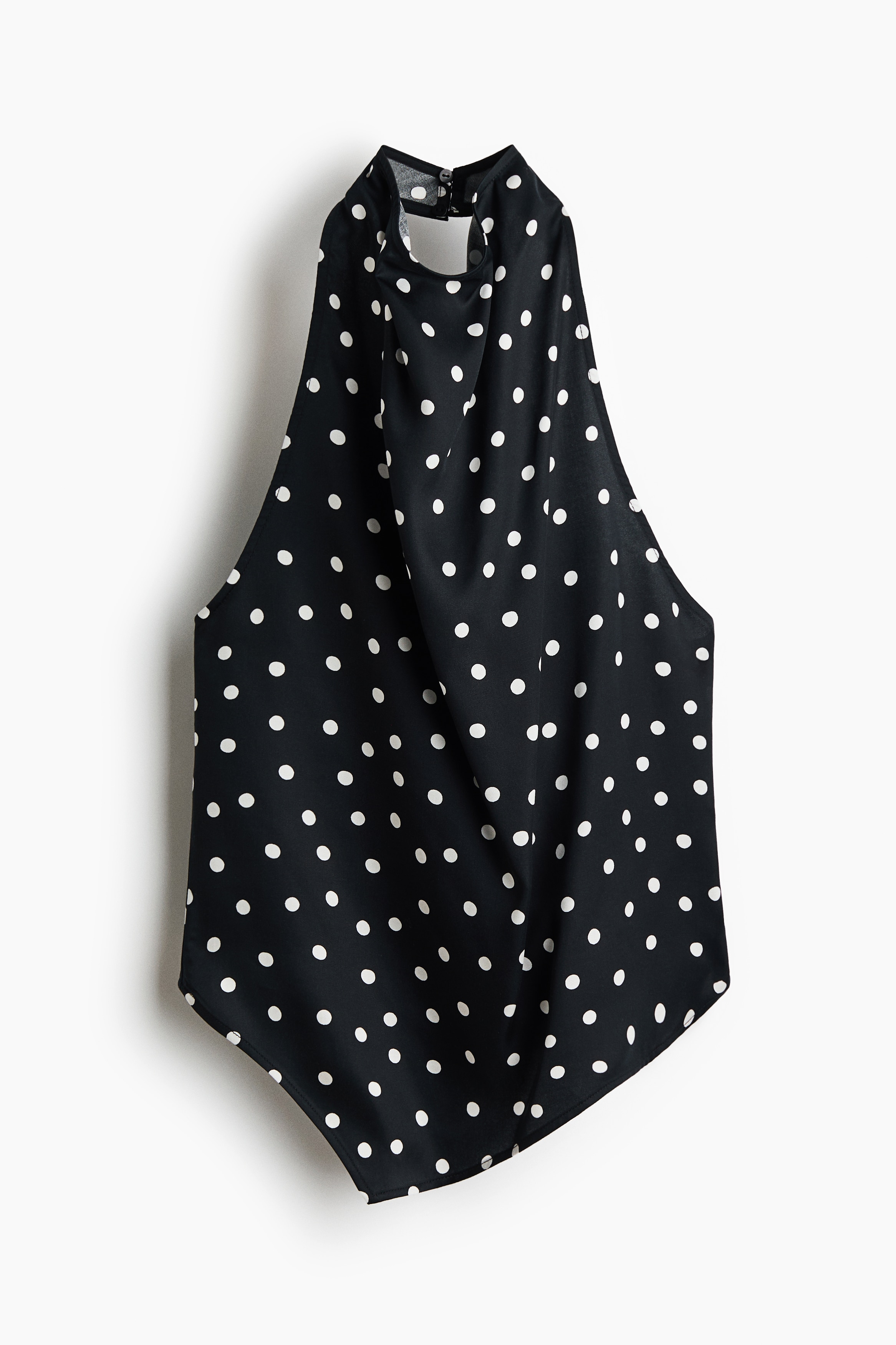 Een zwart satijnen haltertop met witte stippen wordt gedragen door het model, met een gedrapeerde watervalhals en een asymmetrische puntige zoom. De top is gestyled met een zwarte broek, waarin het model beide handen steekt.