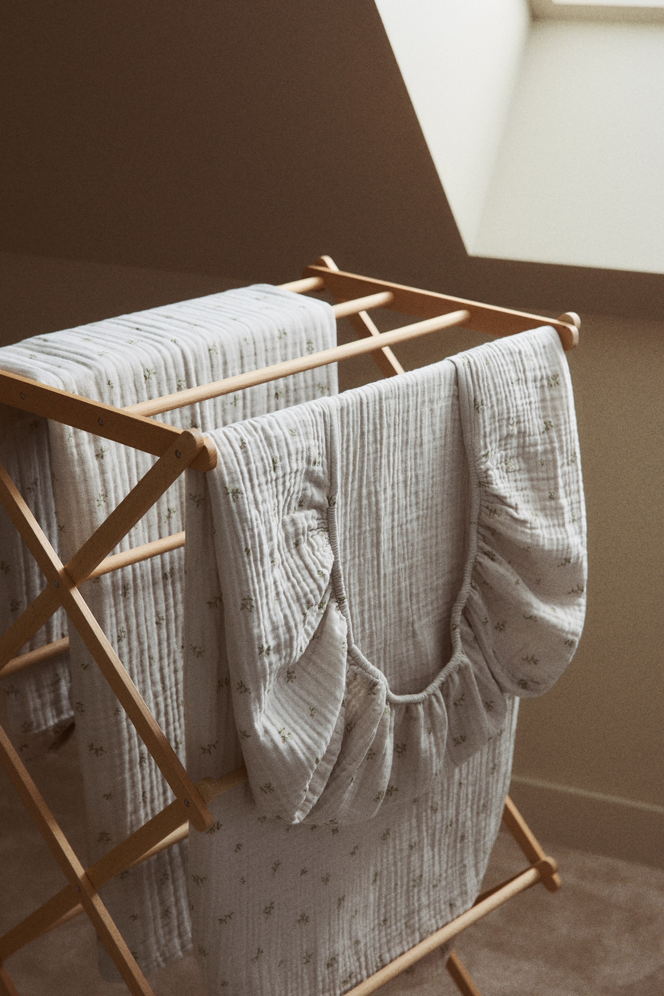 View larger image: A snug cot sheet in gentle cotton muslin is elegantly arranged over a wooden drying rack, showcasing its full print of delicate green leaf motifs and its visibly gathered elasticated edge.