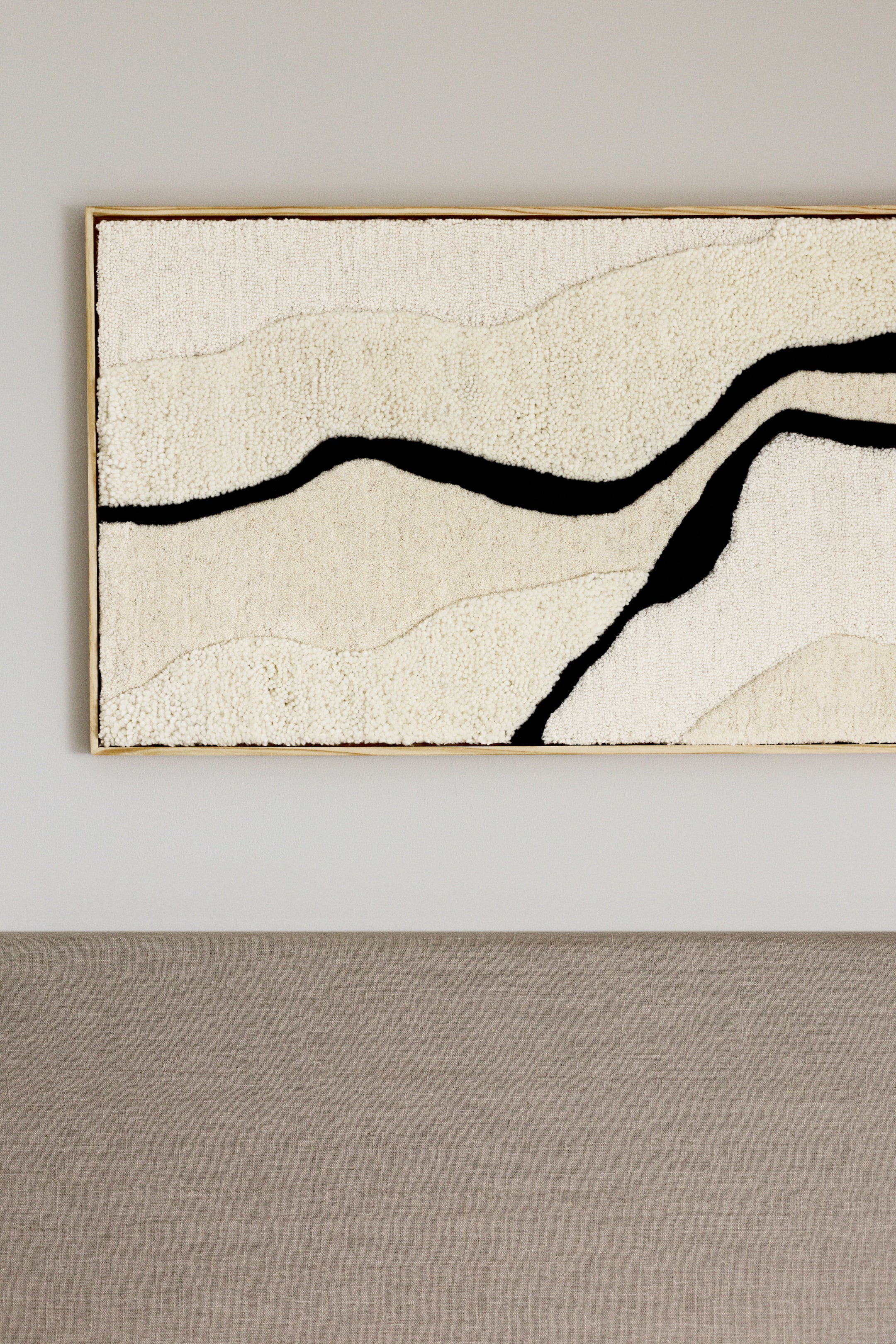View larger image: A rectangular framed textile art piece is displaying an abstract design, featuring undulating black lines against textured fields of off-white and light beige loop pile. It is enclosed within a light wooden frame.