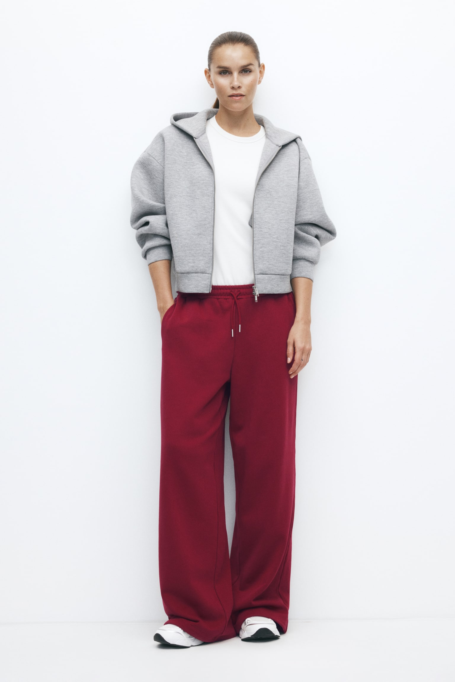 Activewear track pants in a deep red colour, featuring an oversized, wide-leg fit and an elasticated drawstring waistband, are being worn by the model, who is resting one hand in a side pocket. The pants are styled with a white T-shirt, a grey zip-up hoodie, and white trainers. - 1