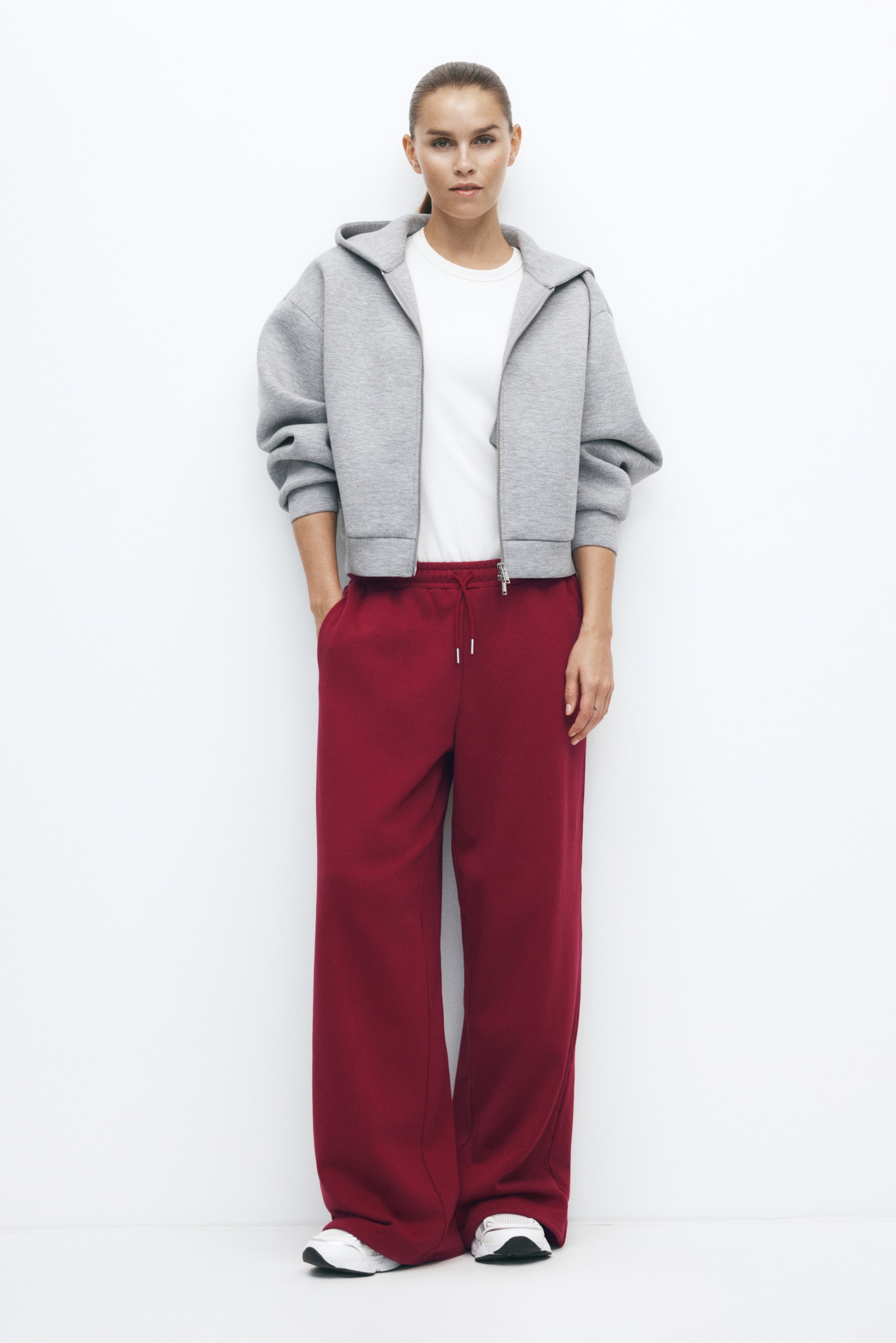 Größeres Bild anzeigen: Activewear-Trainingshose in tiefem Rot mit oversized, weitem Bein und elastischem Bund mit Kordelzug, getragen von dem Model, das eine Hand in der Seitentasche abstützt. Die Hose ist mit einem weißen T-Shirt, einem grauen Zip-Hoodie und weißen Sneakers kombiniert.