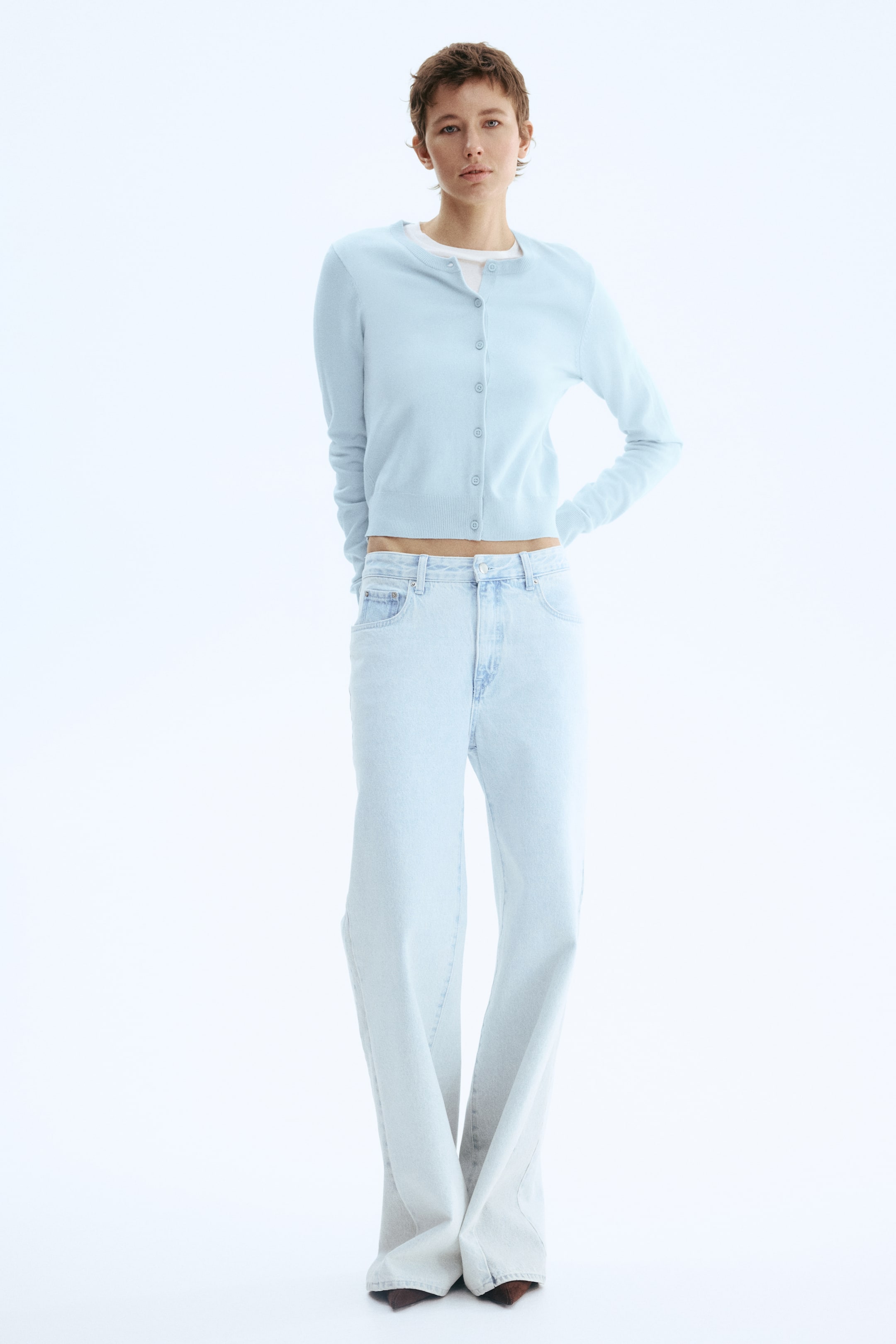 View larger image: The model is donning a light blue fine-knit cardigan, adorned with buttons along the front and ribbing around the neckline, cuffs, and hem. The cardigan is layered over a white top.