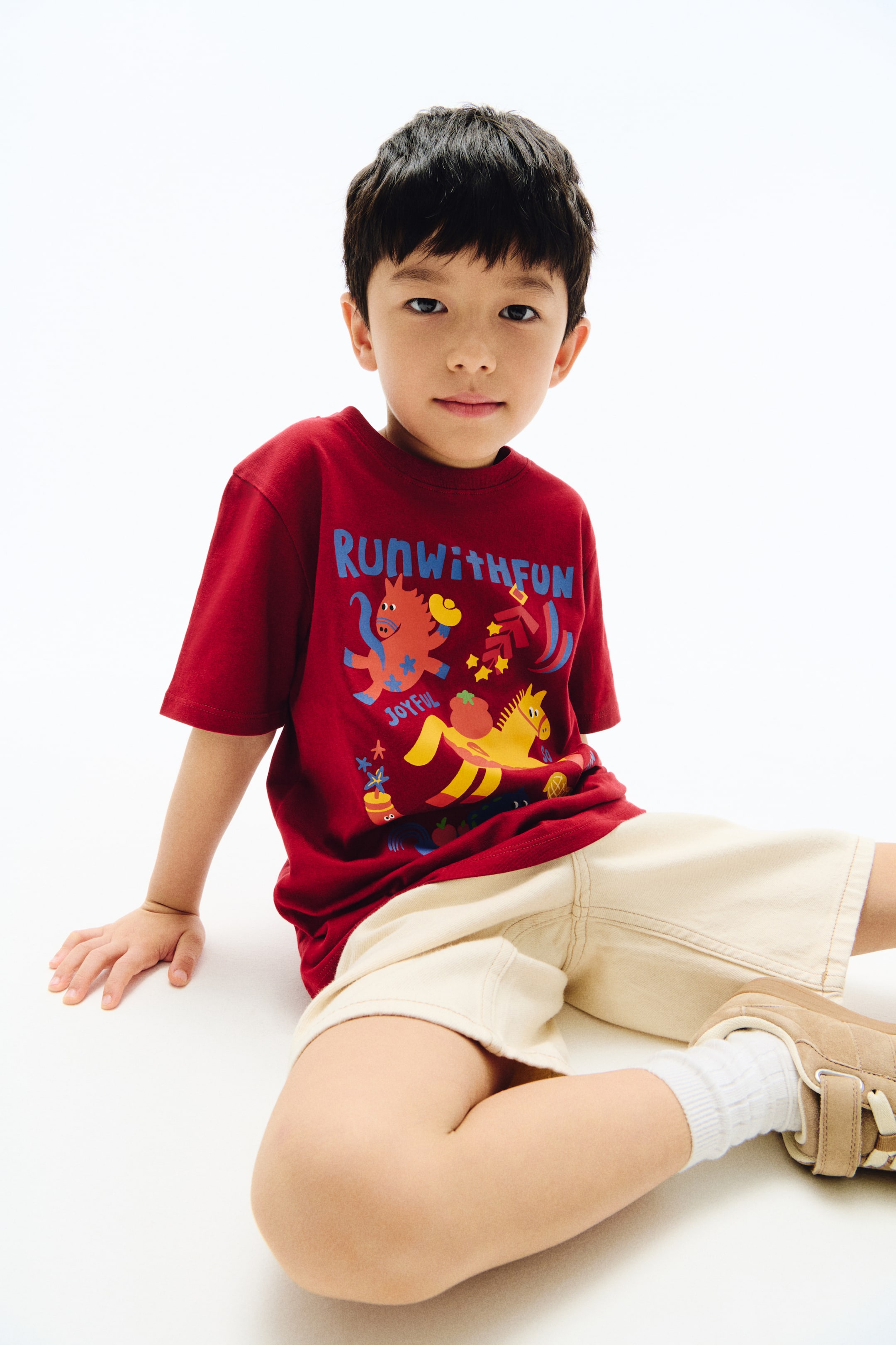 View larger image: A deep red T-shirt featuring a ribbed crew neck and short sleeves. The front showcases a colourful graphic print with blue text reading “RUNWITHFUN” and yellow text reading “JOYFUL”, along with stylised orange and yellow horses, stars, and abstract shapes.
