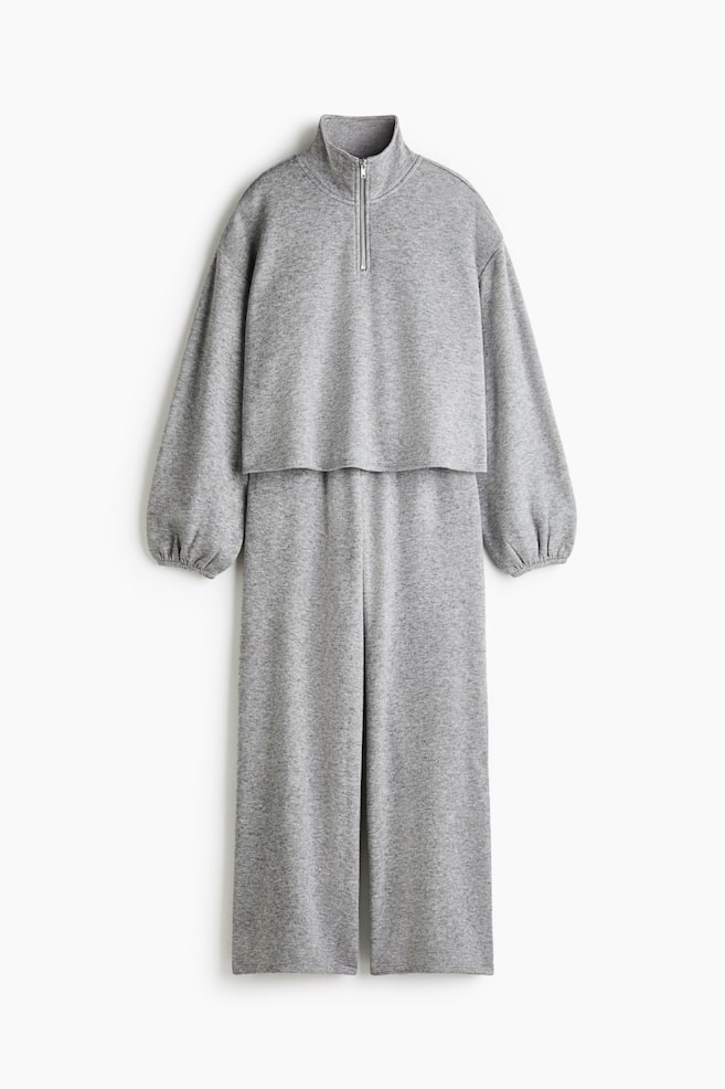 Ein grau melierter Strick-Jumpsuit hängt frontal und verfügt über einen hohen Kragen mit halbem ReiÃverschluss, lange Ballonärmel mit gerafften Bündchen sowie ein kurzes Overlay-Top über einer passenden Hose mit weitem Bein. - 1