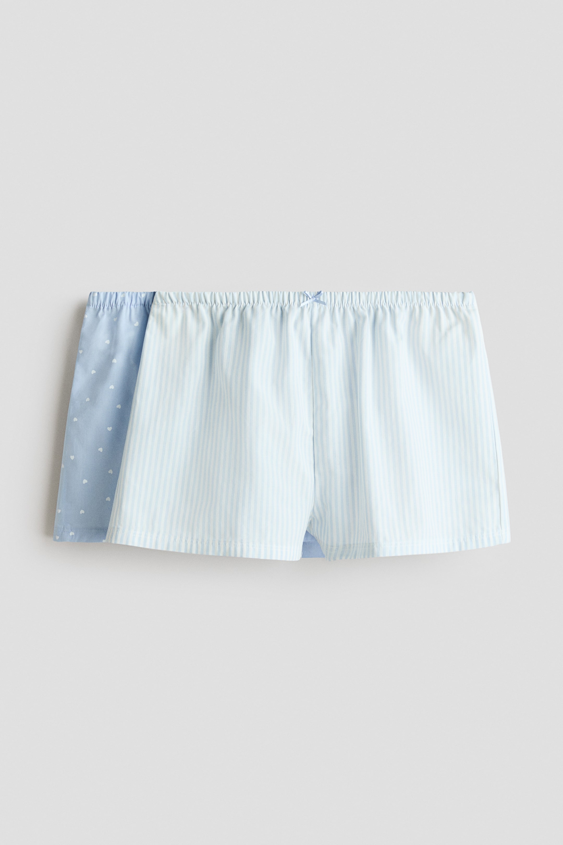 View larger image: Pyjama shorts are shown from the front, with the foremost pair showcasing light blue and white vertical stripes, an elasticated drawstring waist, and a small blue bow at the centre. A second pair of pyjama shorts is placed behind, featuring a light blue fabric adorned with white heart patterns and an elasticated waist.