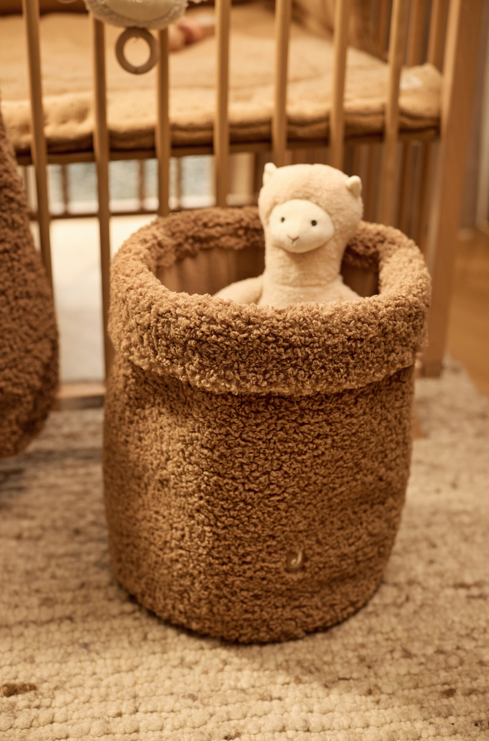 Een cilindervormige opbergmand van gestructureerde teddyfleece in camelbruin staat rechtop en laat een brede omgeslagen rand aan de bovenkant zien. Onderaan aan de voorkant is een klein, vierkant bruin label met een witte letter "J" aangebracht.