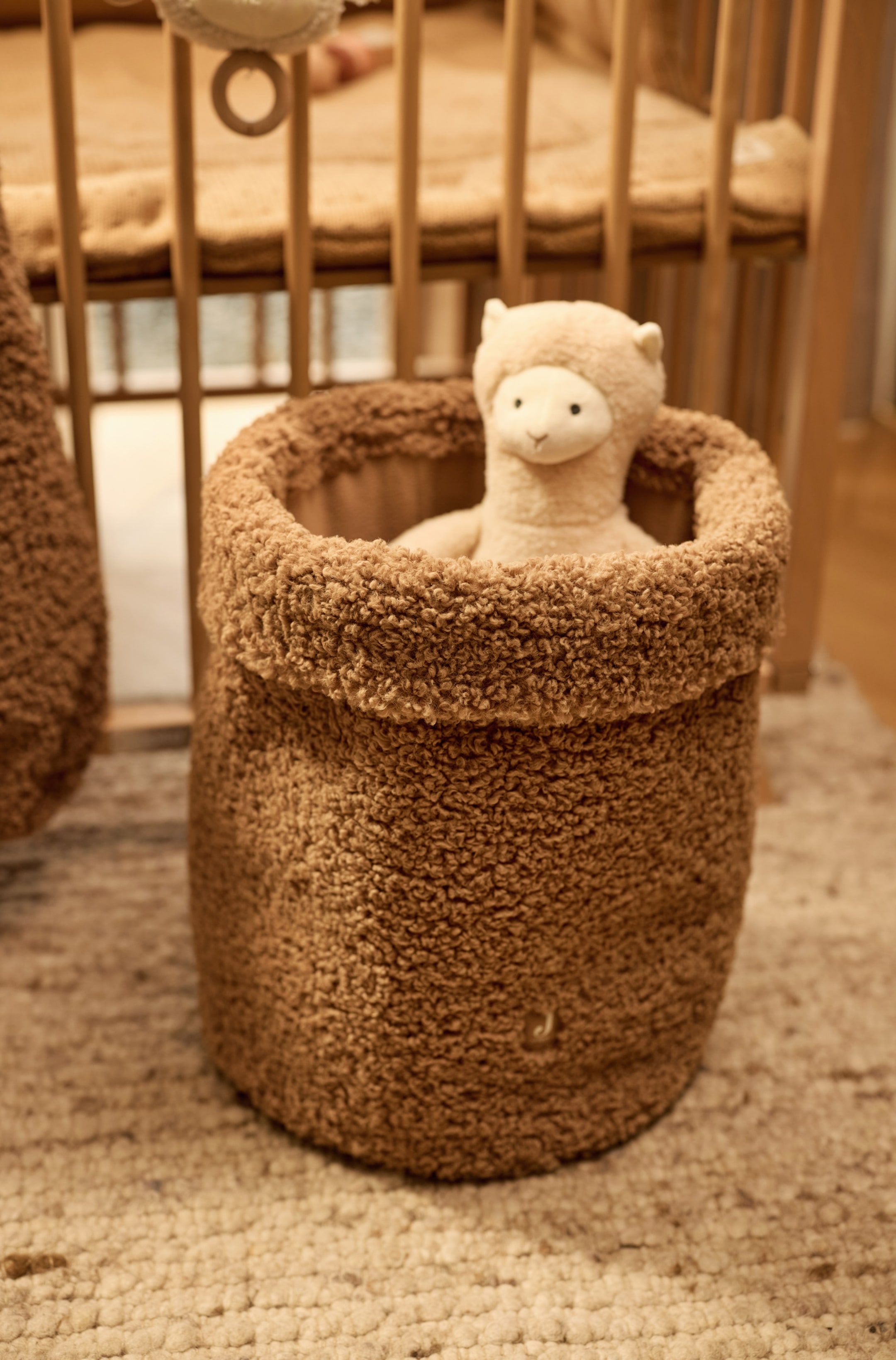 Grotere afbeelding bekijken: Een cilindrische opbergmand van getextureerde, bruine teddyfleece staat rechtop met de bovenrand omgeslagen. Binnenin zit een crèmekleurig knuffellama, dat over de rand heen kijkt.