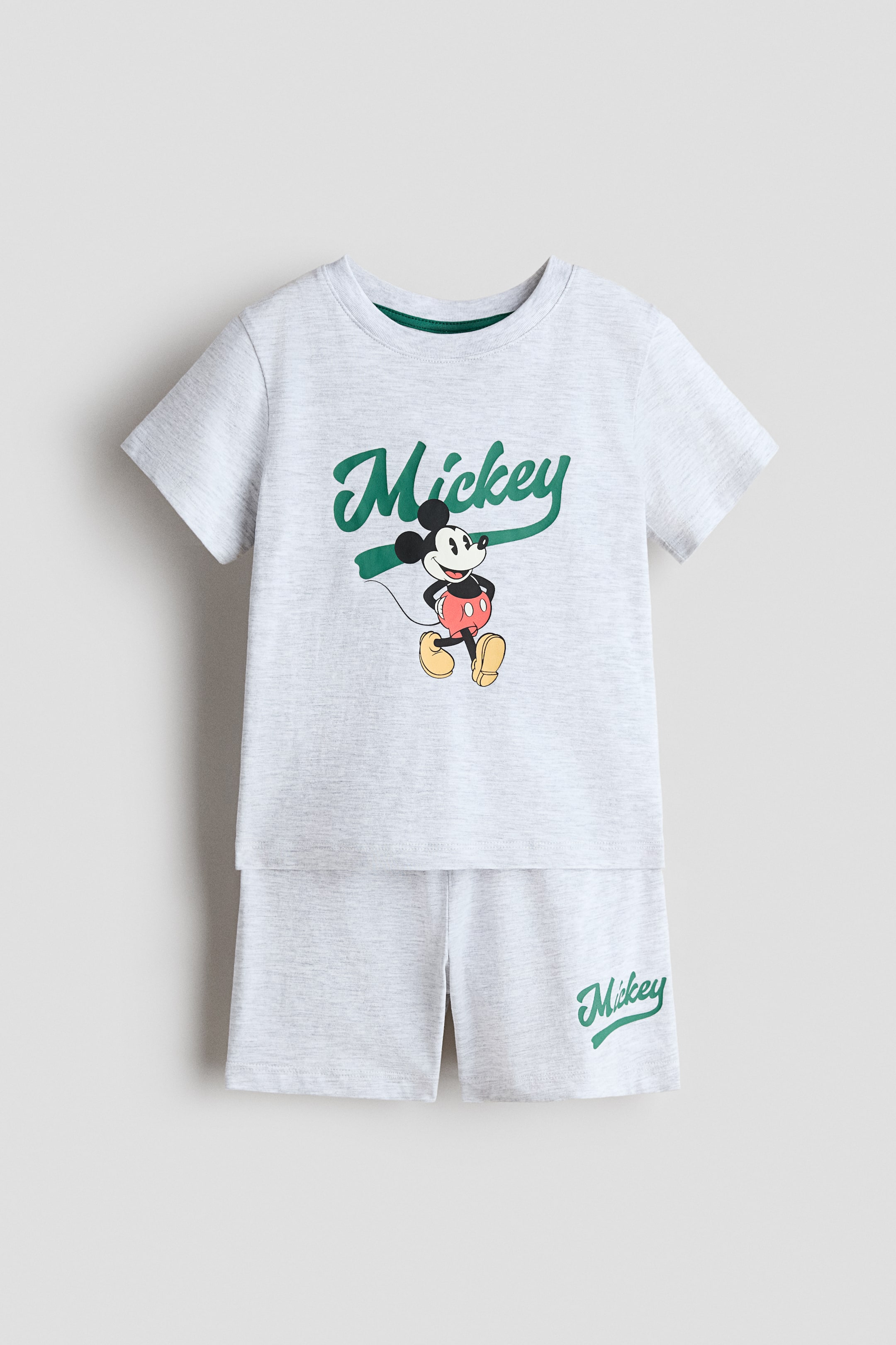 View larger image: A light grey marl pyjama set, crafted from printed cotton jersey, is showcased FRONT, comprising a T-shirt and matching shorts. The T-shirt features a grey ribbed neckline with a dark green inner trim and a green "Mickey" script accompanied by a Mickey Mouse graphic. The shorts boast an elasticated waist and a green "Mickey" script on the left leg.