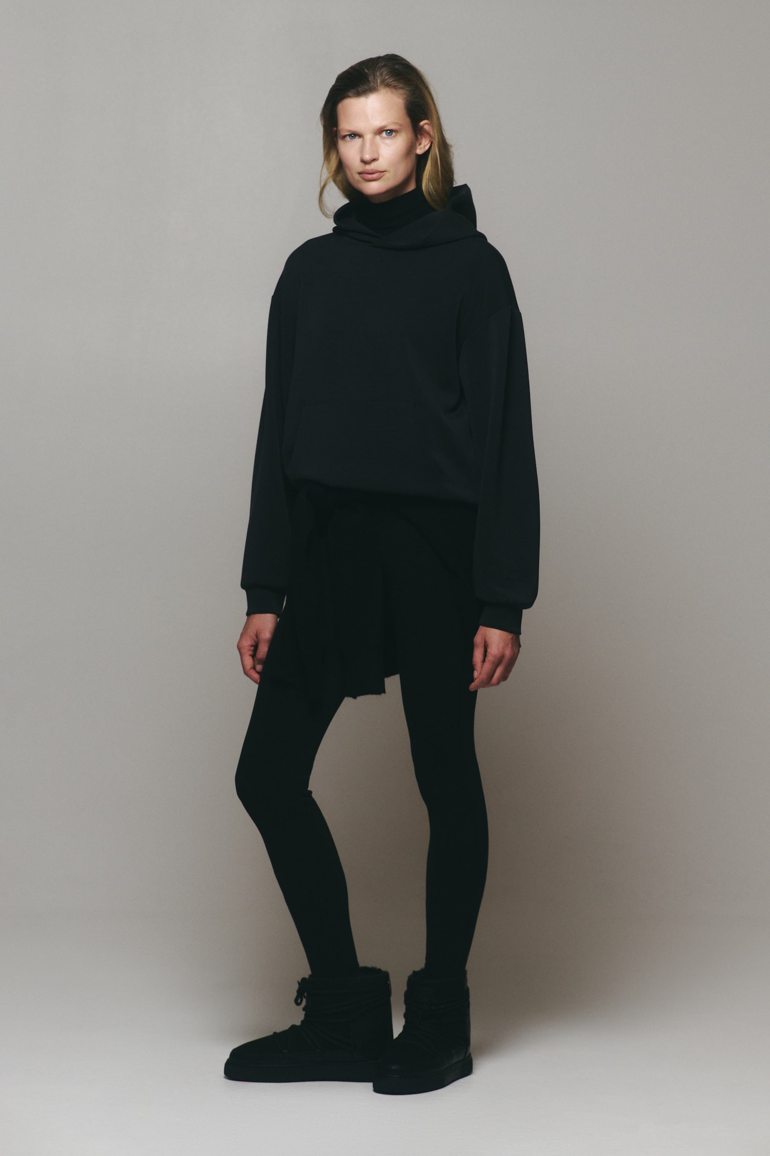 An oversized, dark grey activewear hoodie featuring a lined wrap hood, dropped shoulders, and a front kangaroo pocket is being worn by the model. The hoodie is layered over a black high-neck top and styled with black leggings. - 1