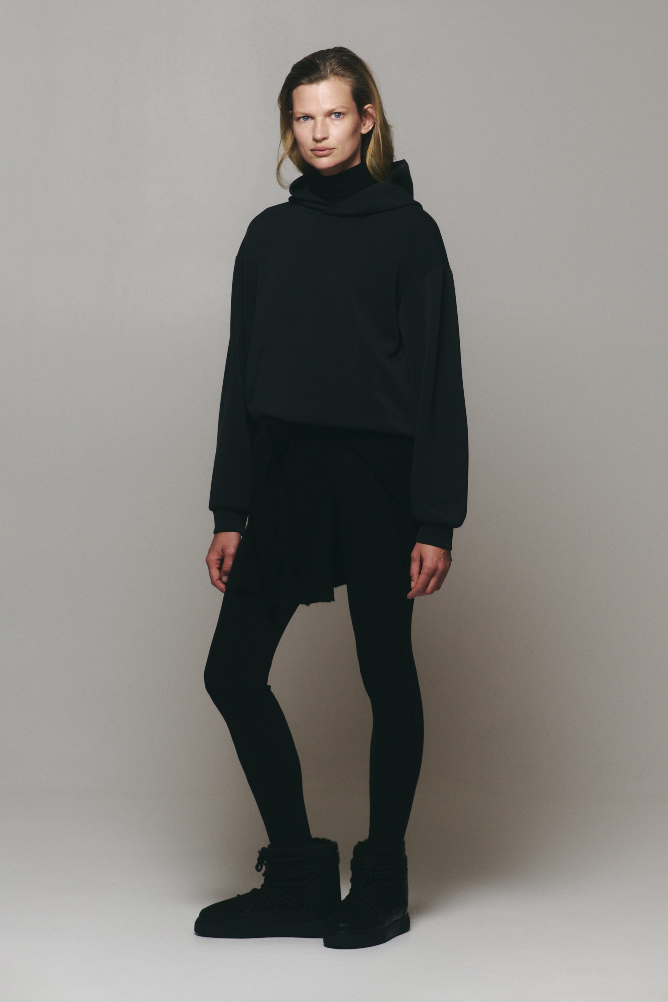 Zobraziť väčší obrázok: Model má na sebe oversized tmavosivú mikinu s kapucňou v aktívnom štýle, s podšívanou obtočenou kapucňou, spadnutými ramenami a predným klokankovým vreckom. Mikina je vrstvená cez čierny top s vysokým golierom a skombinovaná s čiernymi legínami.
