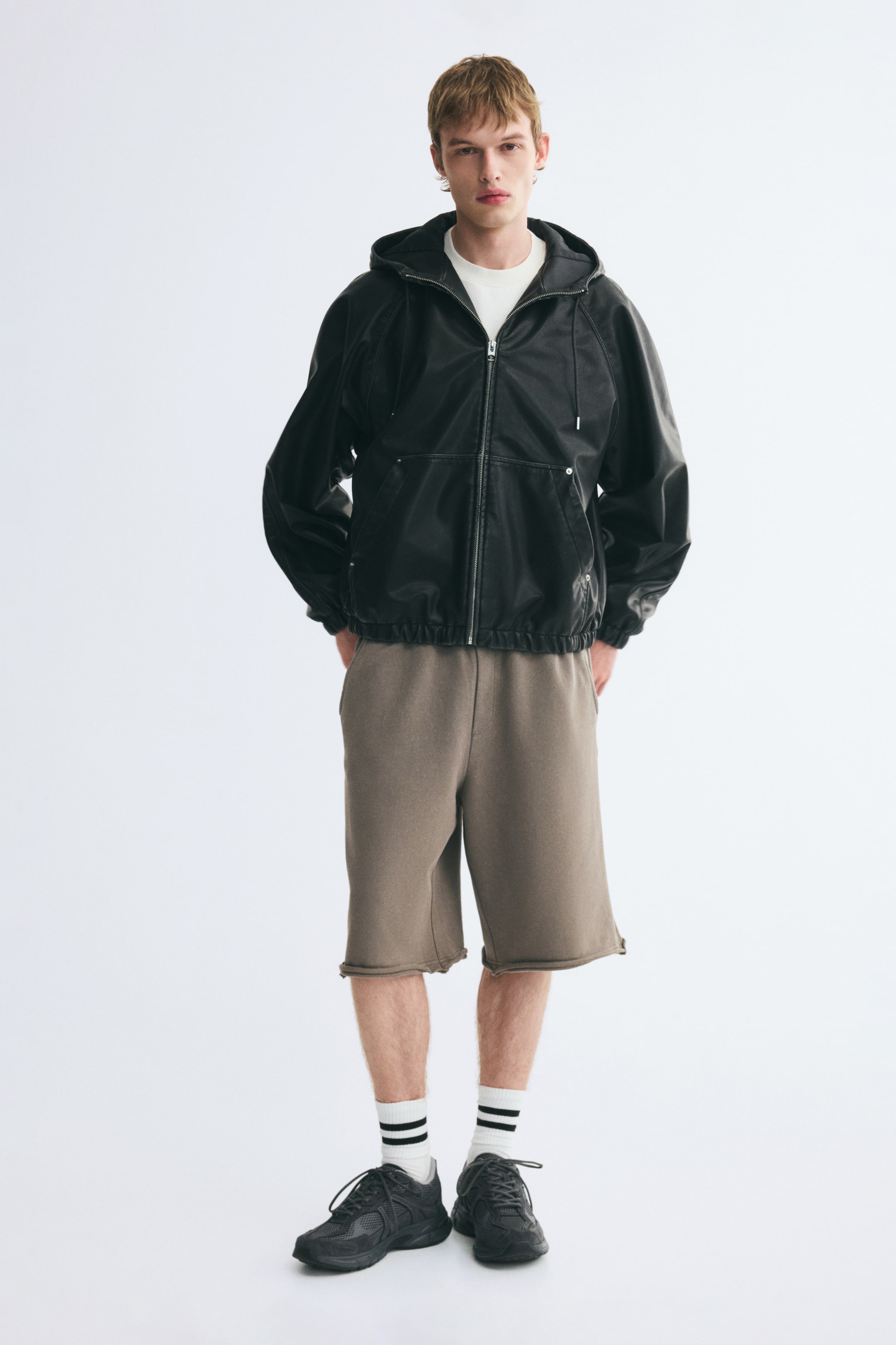 View larger image: A black faux-leather hooded jacket featuring a front zip closure, a drawstring hood, and an elasticated hem is being worn by the model. The jacket is layered over a white t-shirt and styled with brown knit shorts and black sneakers.