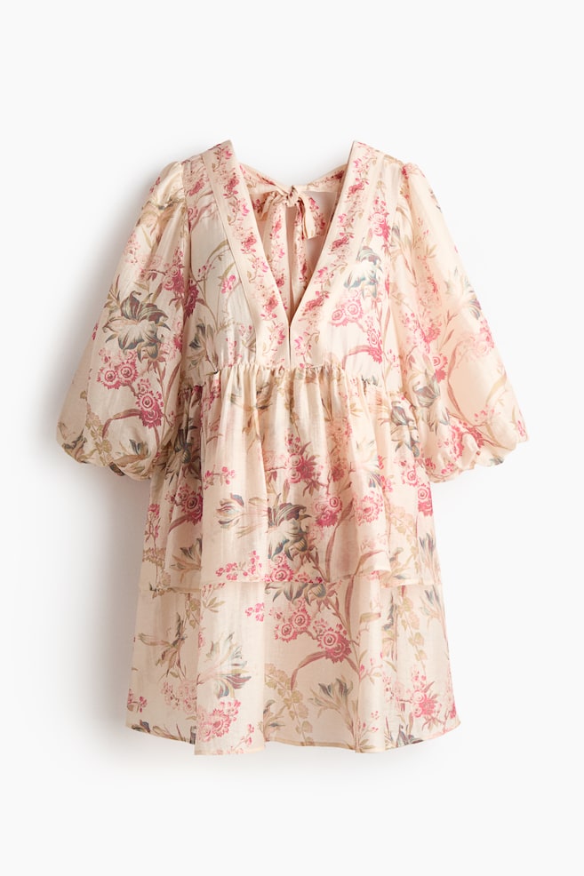 Un vestido color crema claro presenta un vibrante estampado floral en tonos rosa, rojo y verde y está diseñado con un pronunciado escote en V. Además, cuenta con un cierre con lazada visible en la parte posterior del cuello, mangas abullonadas de largo tres cuartos recogidas en los puños y una línea de talle imperio que fluye hacia una falda a capas. - 1