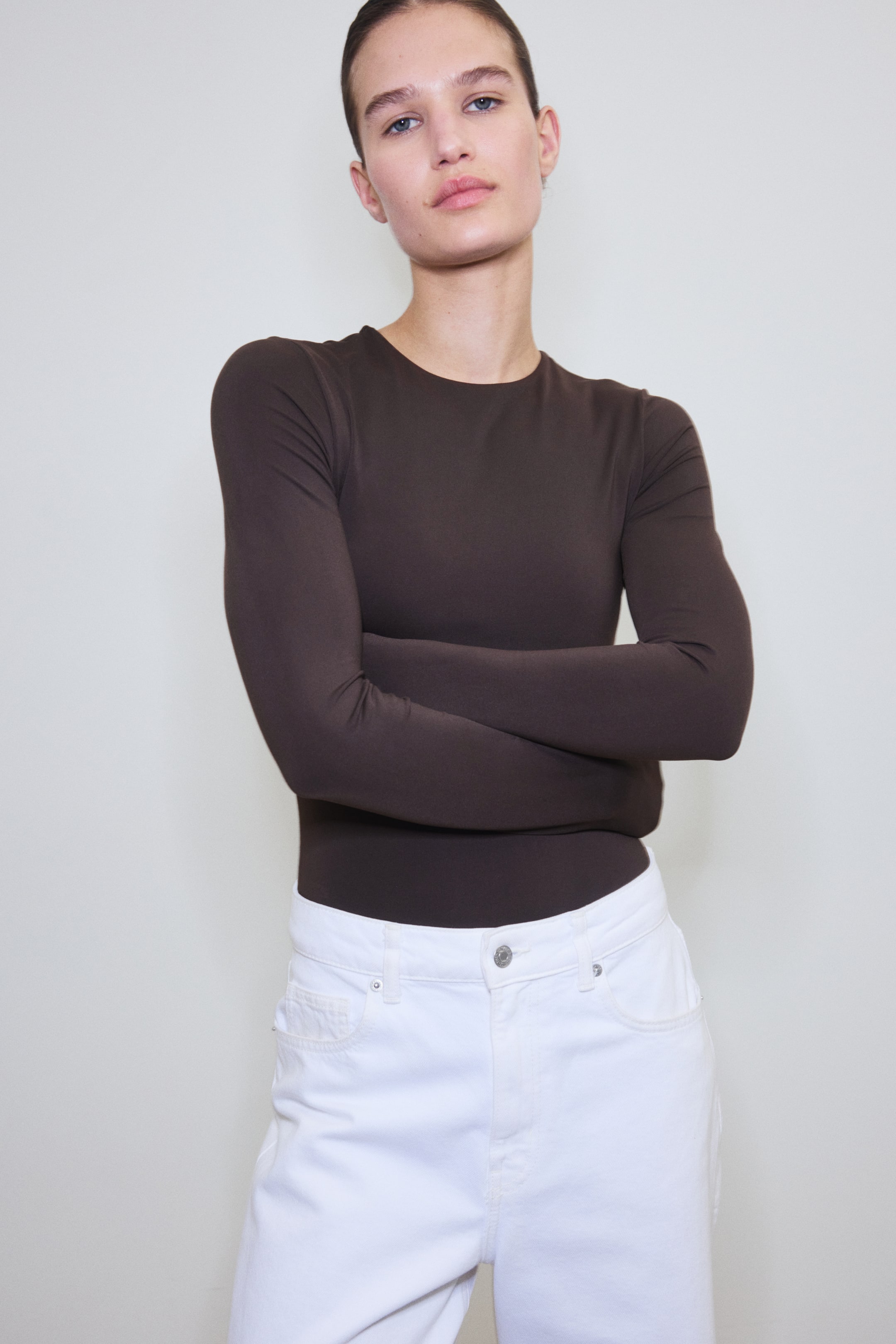 Se større billede: En langærmet mørkebrun bodysuit med rund halsudskæring bæres af modellen. Modellen kombinerer den med hvide bukser.