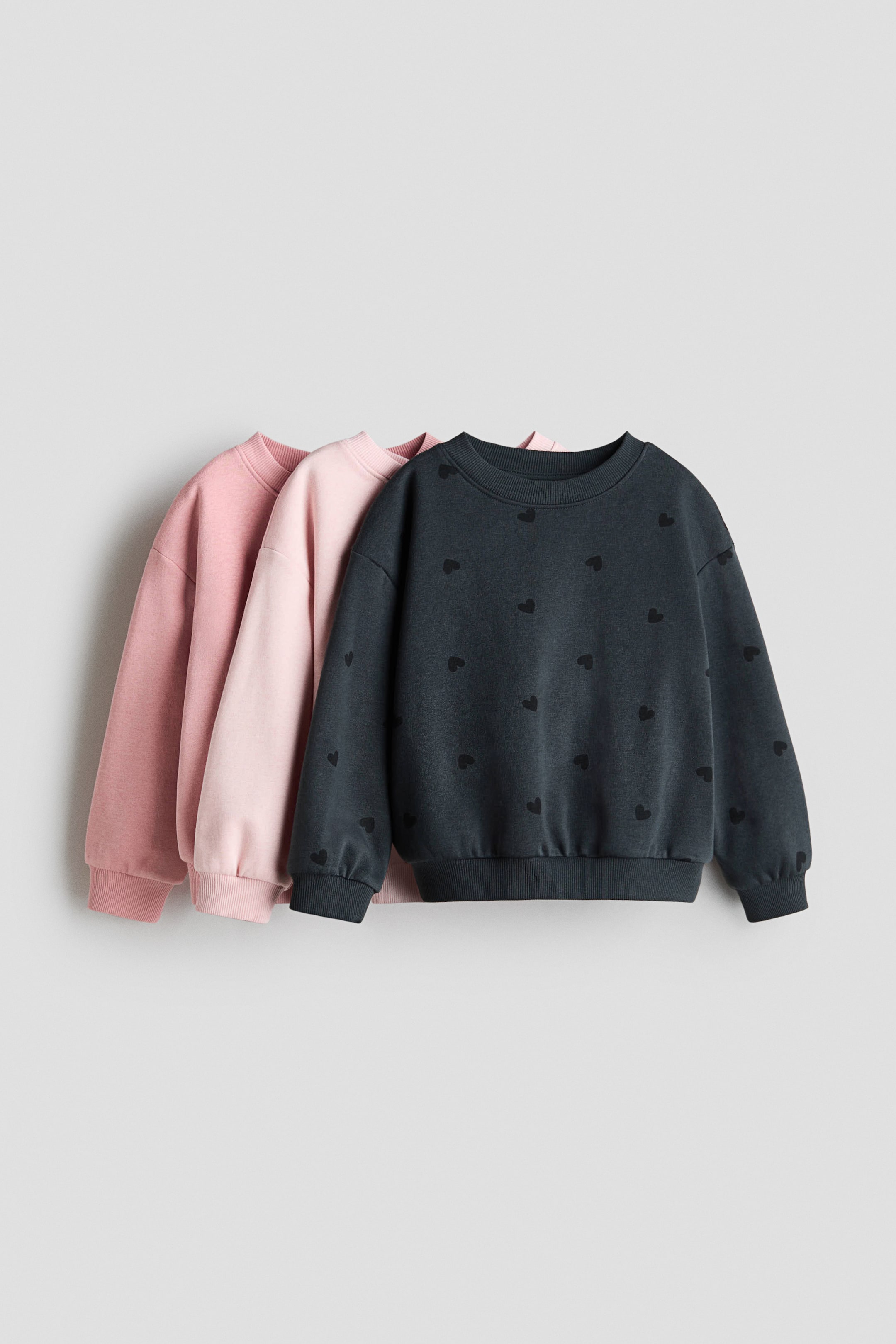 View larger image: Three crew neck sweatshirts are layered, facing forward at a slight angle. The dark grey front sweatshirt is displaying a repeating heart pattern, ribbed cuffs, hem, and crew neck, while two solid pink sweatshirts are visible behind it, also featuring ribbed cuffs, hems, and crew necks.