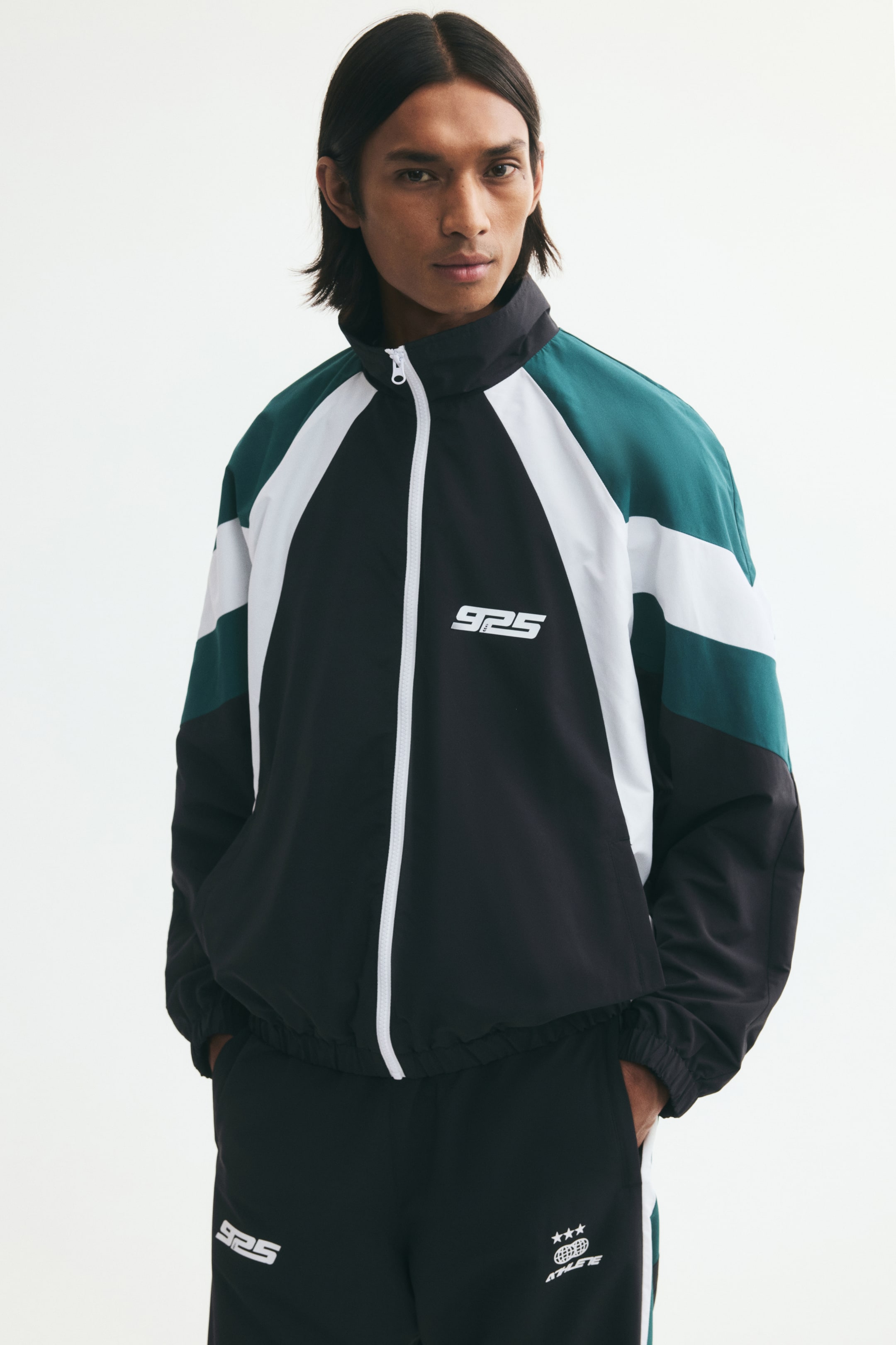 View larger image: A colour-blocked track jacket in black, white, and dark green is being worn by the model. The jacket features a stand collar, a white front zipper, and a small white logo on the chest, and is paired with matching black track pants.