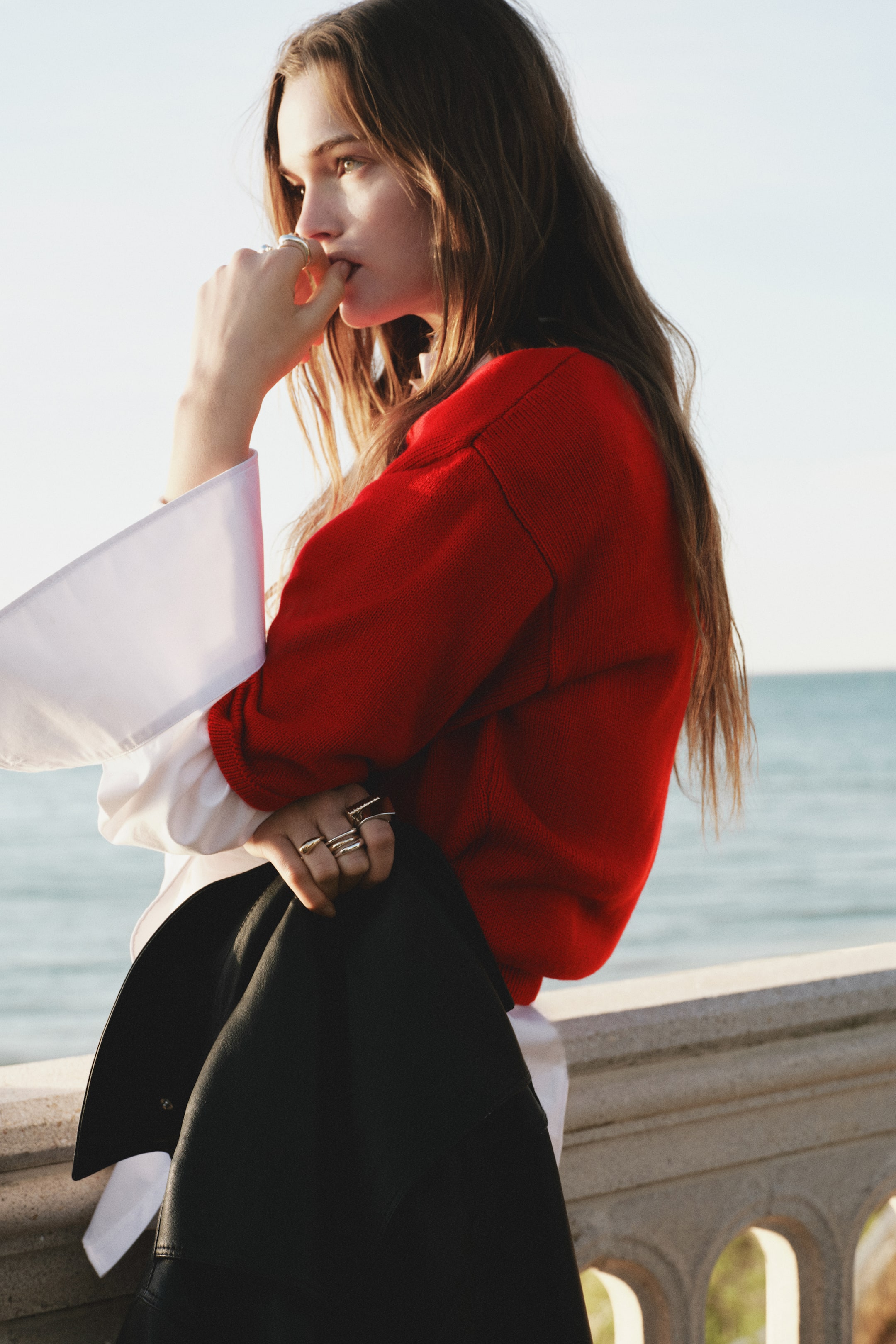 View larger image: A red knitted jumper showcases a relaxed fit with long sleeves, worn layered over a white shirt. The wide cuffs of the white shirt extend beyond the jumper's sleeves.