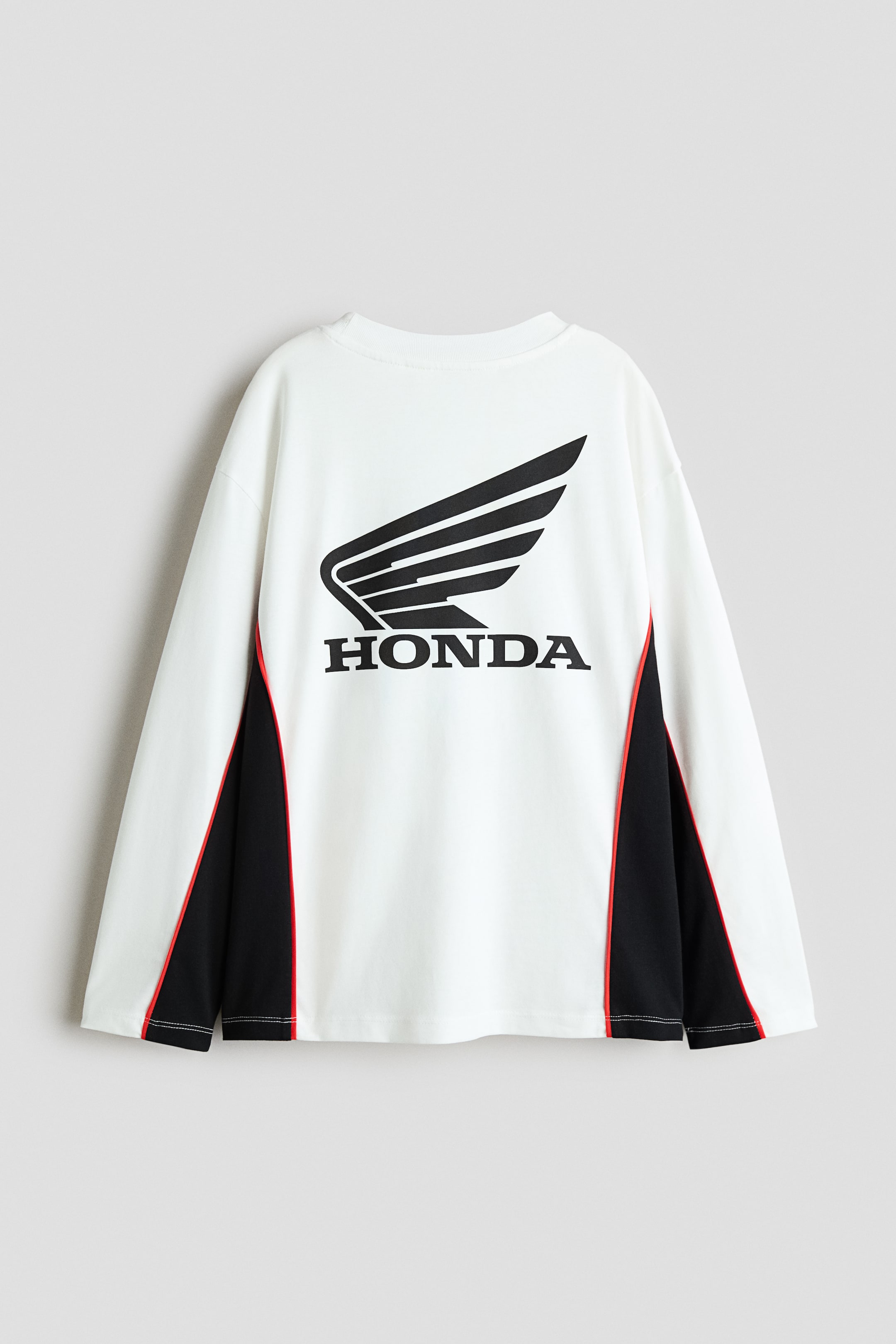 Agrandir l'image: Un t-shirt blanc à manches longues et à col rond est présenté de dos, avec un grand logo Honda ailé noir et du texte imprimés au centre. Le haut intègre des empiècements noirs sur les côtés inférieurs et sous les aisselles, soulignés par un fin passepoil rouge.