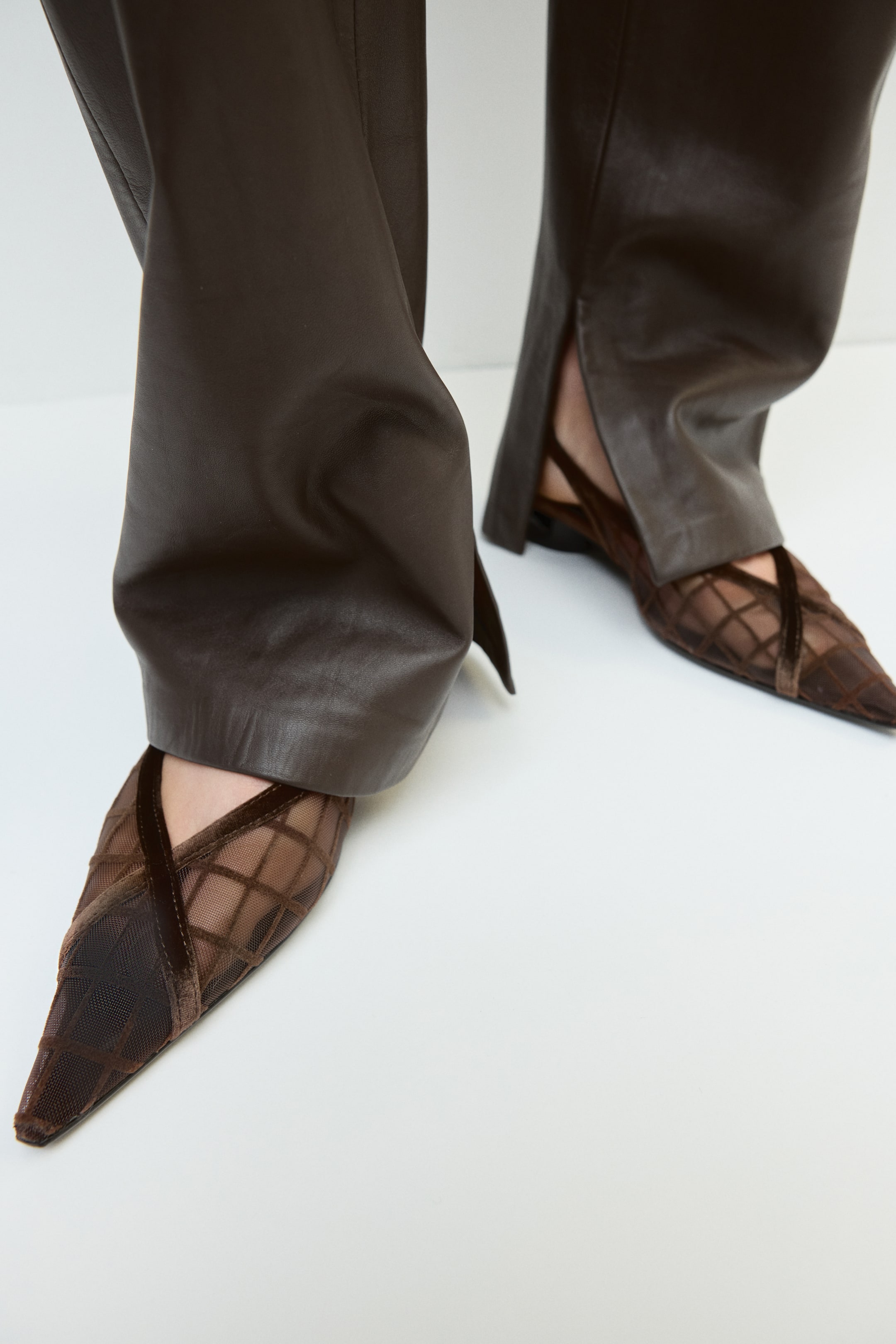 Ver imagen más grande: La modelo lleva unos zapatos slingback de tacón ancho en malla marrón y terciopelo, con punteras afiladas y tiras decorativas de terciopelo cruzadas, además de un diseño de rejilla. Los zapatos se combinan con unos pantalones de efecto piel en marrón oscuro con bajos abiertos en los tobillos.