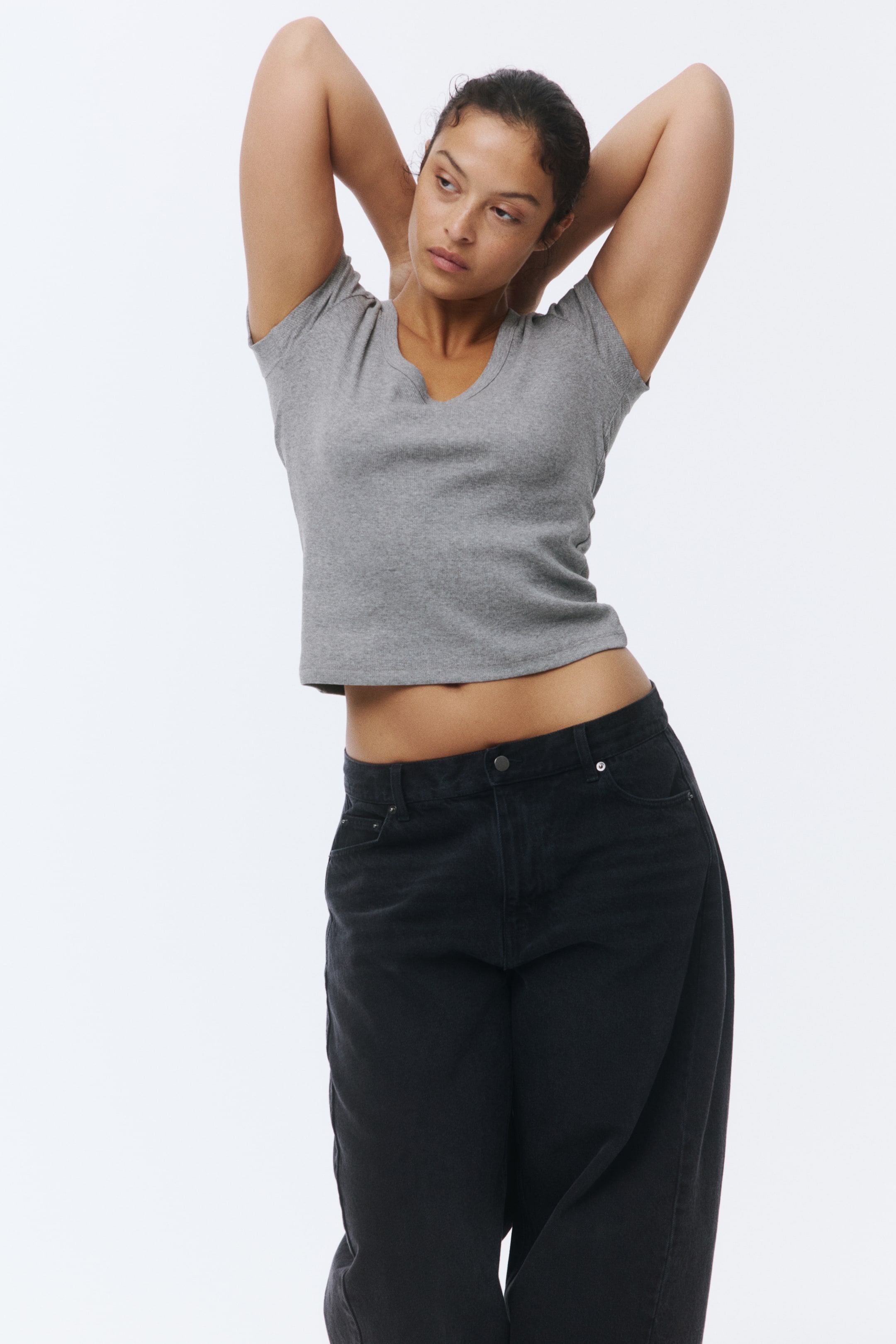 View larger image: The model is wearing a fitted grey ribbed cotton T-shirt featuring a V-neckline and a straight hem. The T-shirt is cropped, exposing the model's midriff, and she is raising her arms.