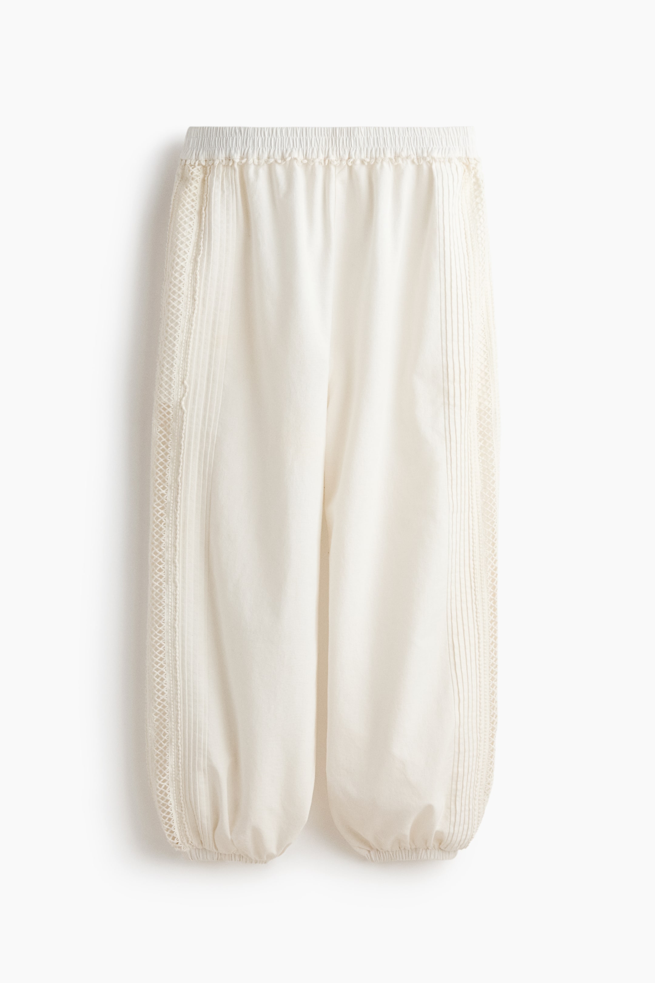View larger image: The cream-coloured trousers are displayed from the front, highlighting an elasticated and ruched waistband. Vertical lace trim runs alongside pintuck details down the sides of the legs, which gather into elasticated cuffs at the ankles.