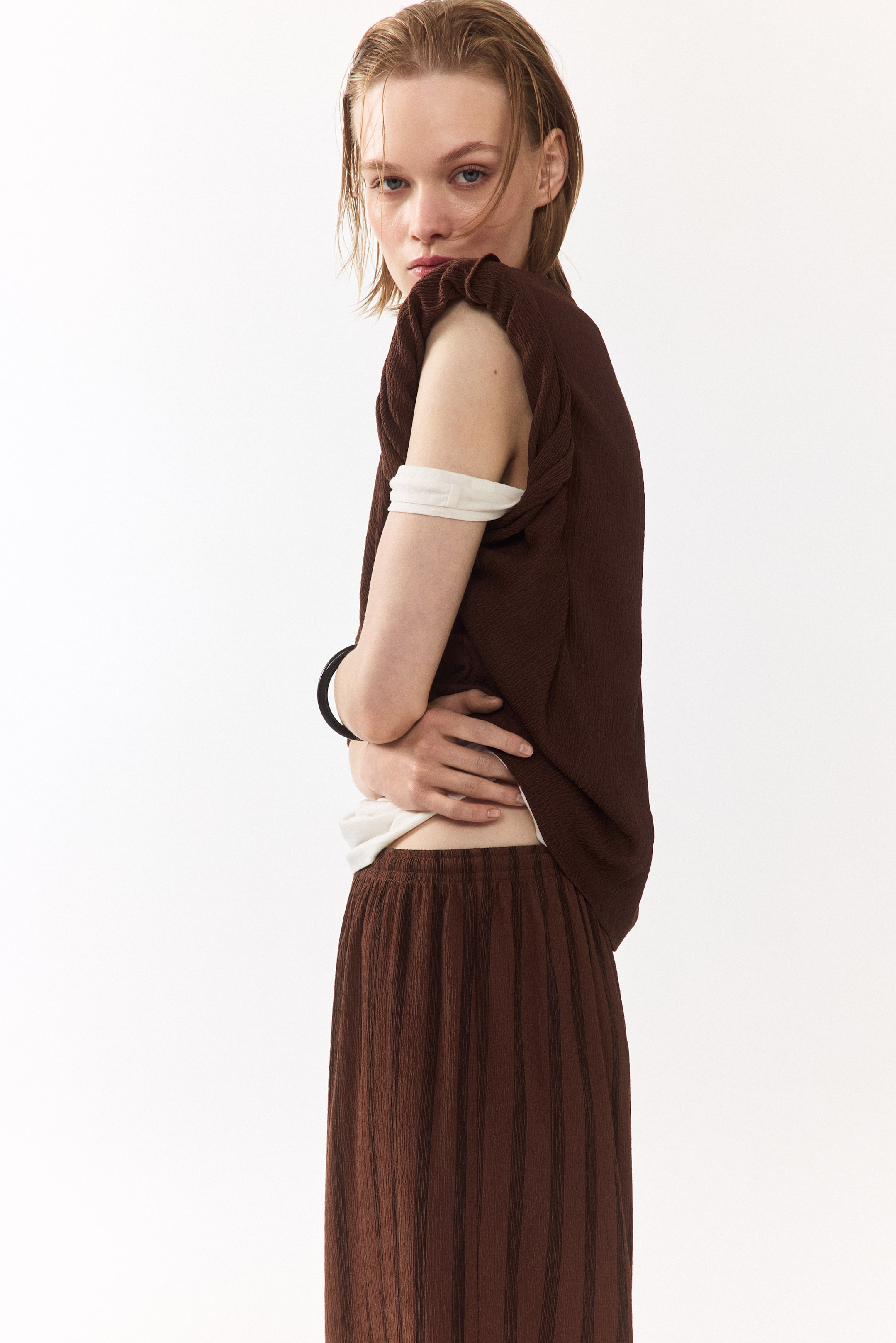 Voir une image plus grande: Un haut sans manches texturé brun est porté par-dessus un haut à manches longues de couleur claire, avec des emmanchures froncées. Un pantalon large texturé brun assorti est également visible, présentant une taille élastique et des rayures verticales subtiles.