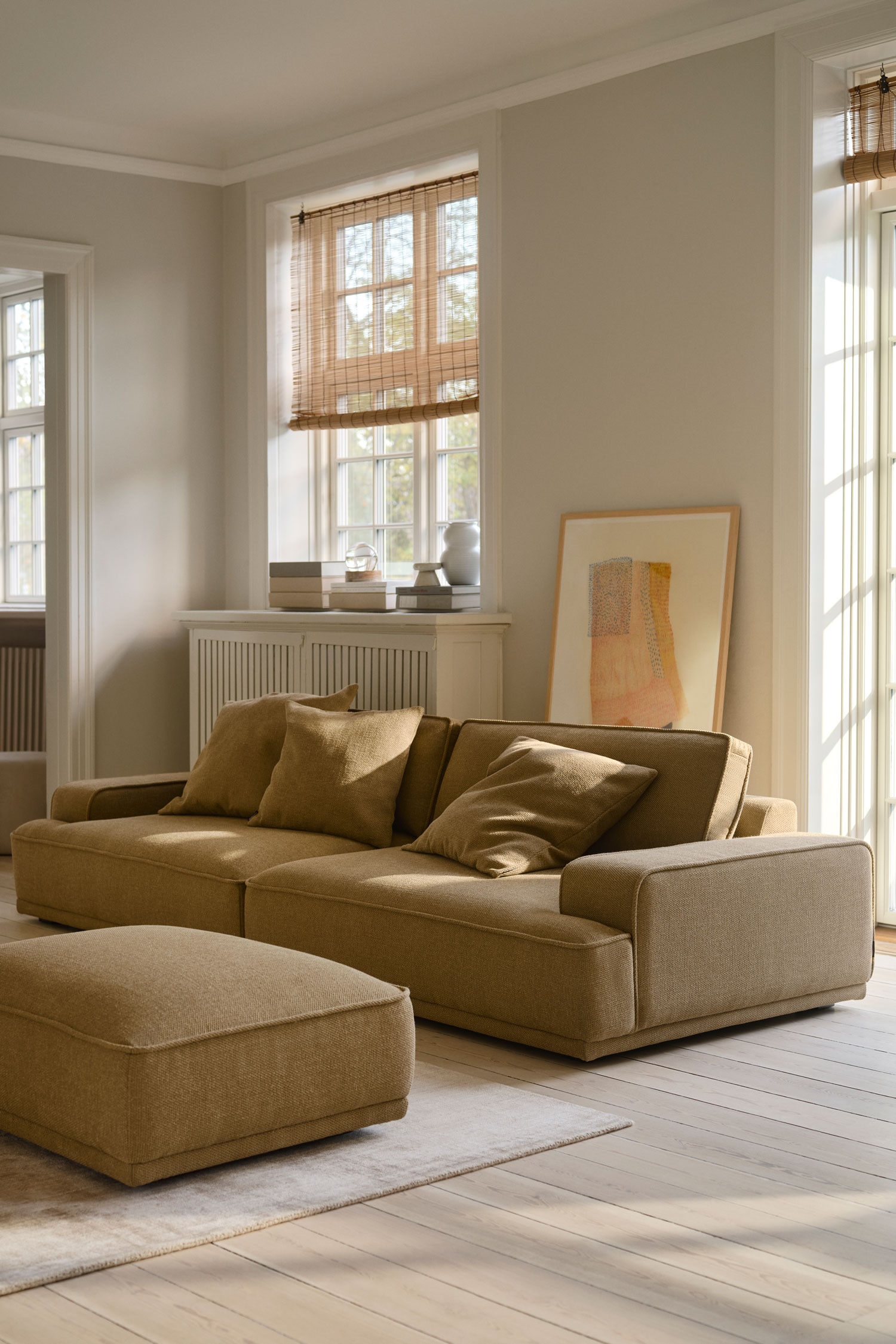 Een rechthoekige gestoffeerde ottoman in een gestructureerde beige stof staat naar voren gericht en rust op lage, donkere blokpoten. Hij heeft een licht gewelfd zitkussen met afgewerkte naaddetails en een laag, hoekig silhouet.