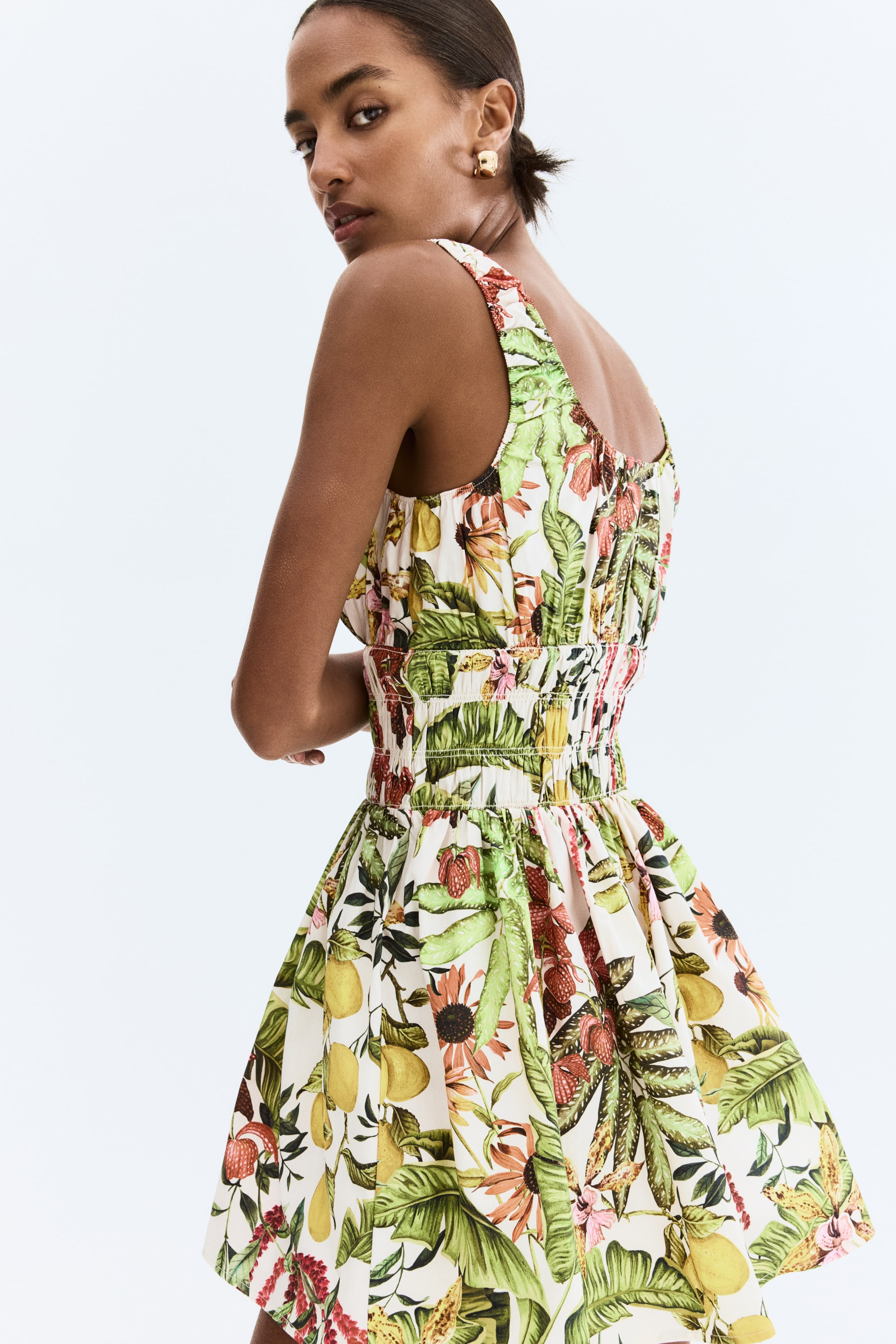 Ampliar la imagen: El vestido presenta un estampado tropical de hojas verdes, limones amarillos y fresas rojas sobre un fondo blanco. Exhibe tirantes anchos, una cintura fruncida y una falda completa y acampanada, presentado desde un ángulo trasero.